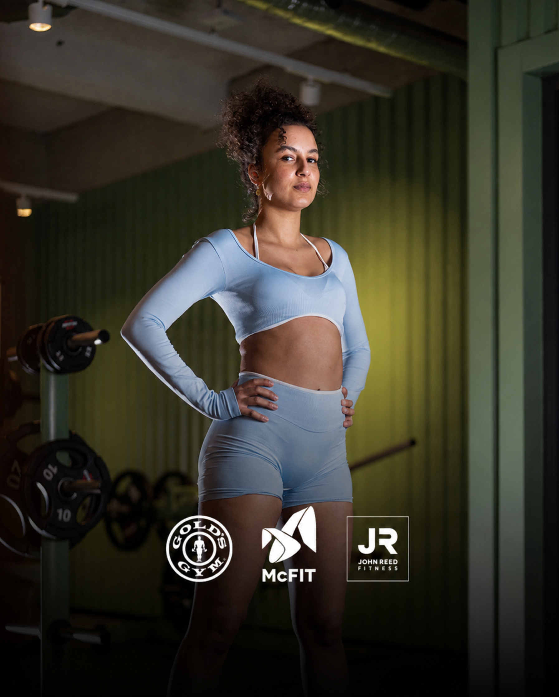 Eine junge Frau in Pastell-Blau Workout-Set steht in einem Fitnessstudio mit Gewichtsstange im Hintergrund, Blick selbstbewusst.