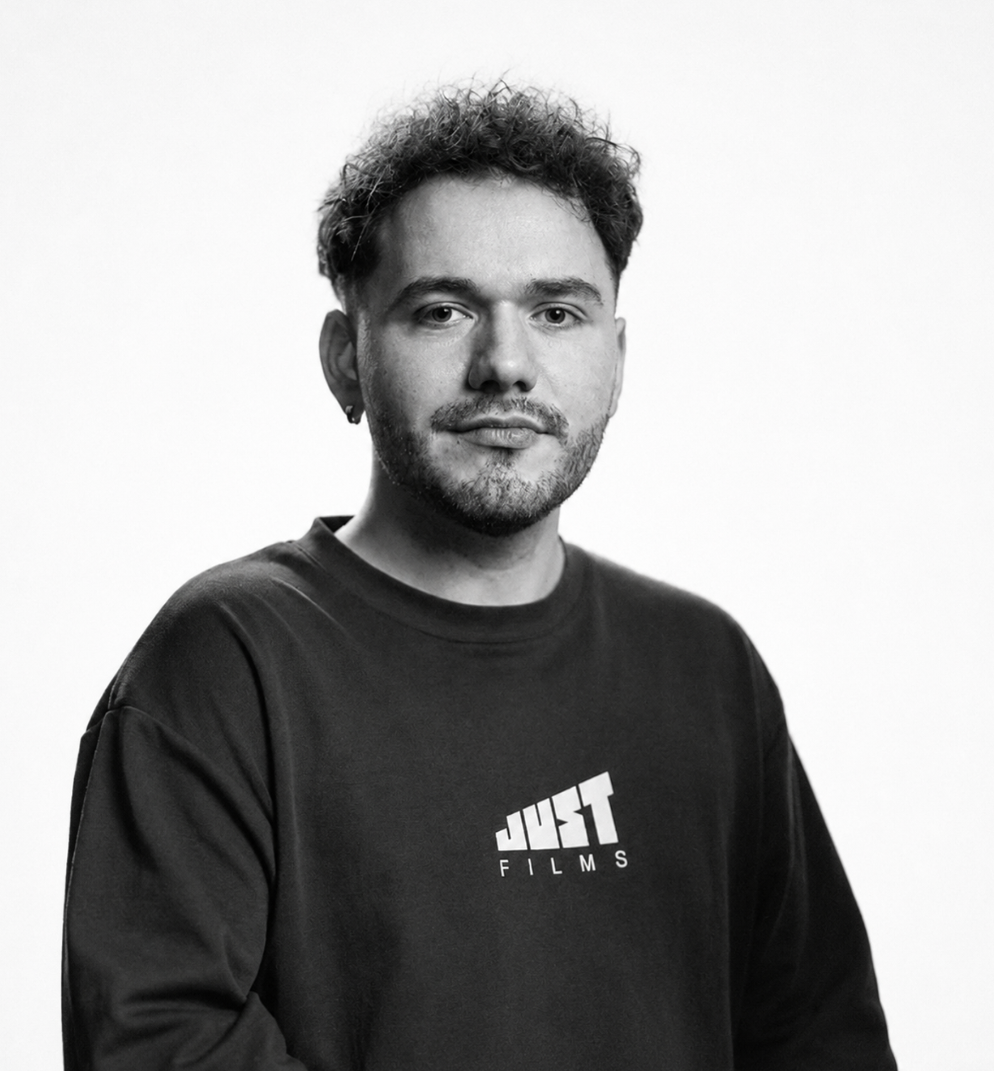 Porträt eines jungen Mannes mit dunklem Kurzhaarschnitt, grau gesträhntem Haar und E-Inseln, trägt ein schwarzes T-Shirt mit weißem Text. Hintergrund ist weiß.