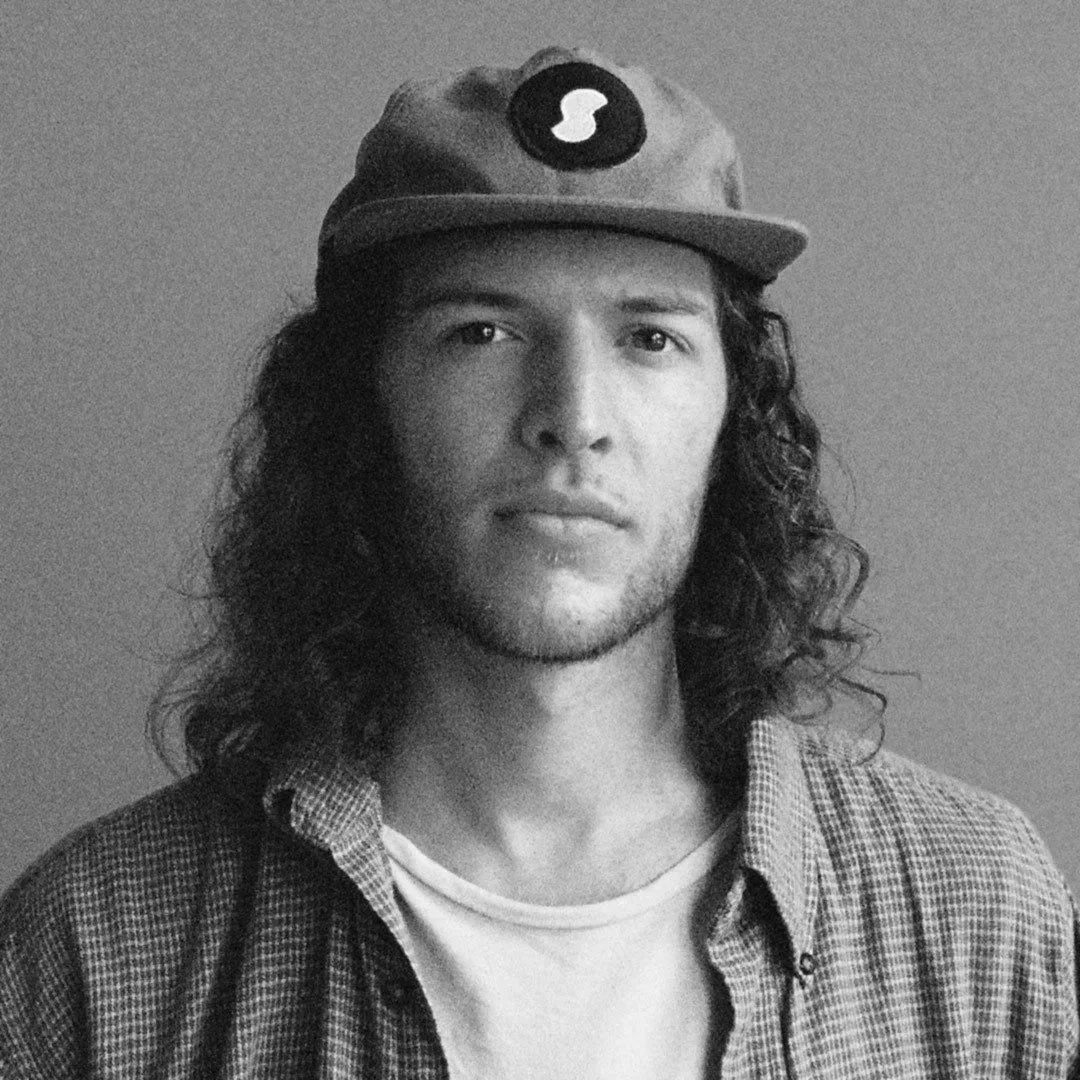 Black and white photo of a man with long, curly hair wearing a baseball cap and a checkered shirt.