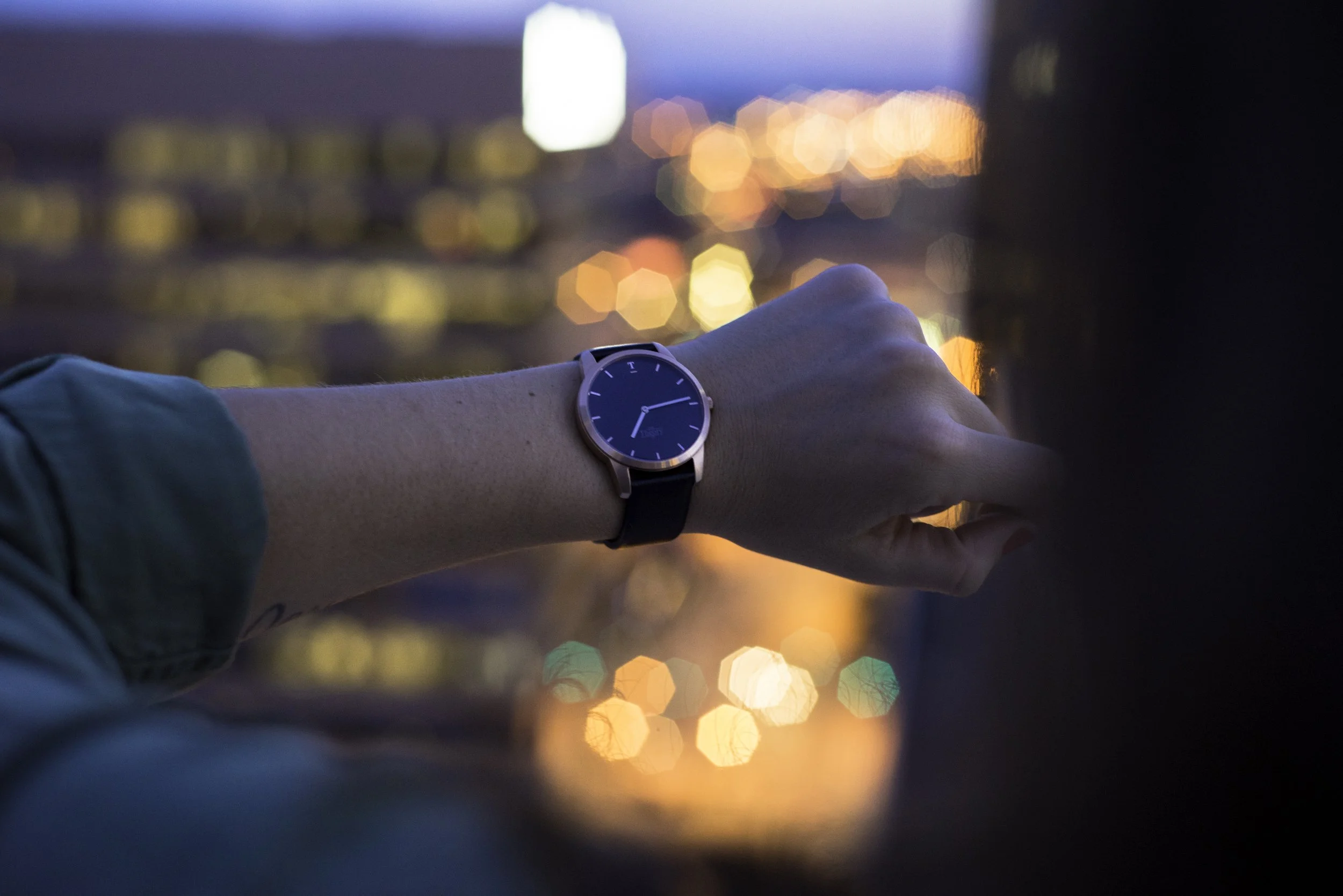 A person's left wrist with a smartwatch showing the time 9:10. The background shows a city skyline at dusk with colorful bokeh lights.