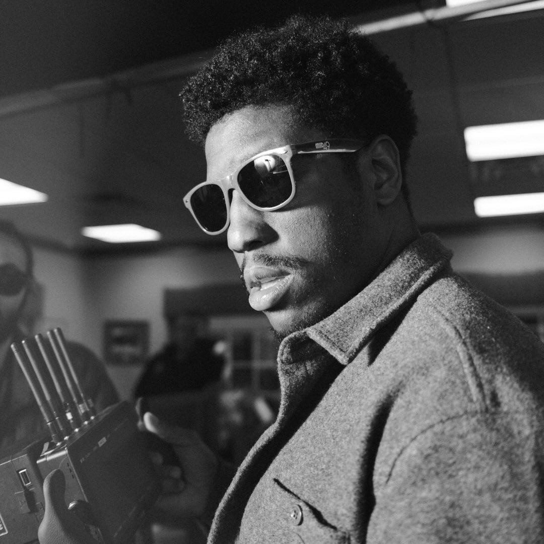 A young man with curly hair wearing sunglasses and a coat, holding a camera, in a black and white indoor setting.