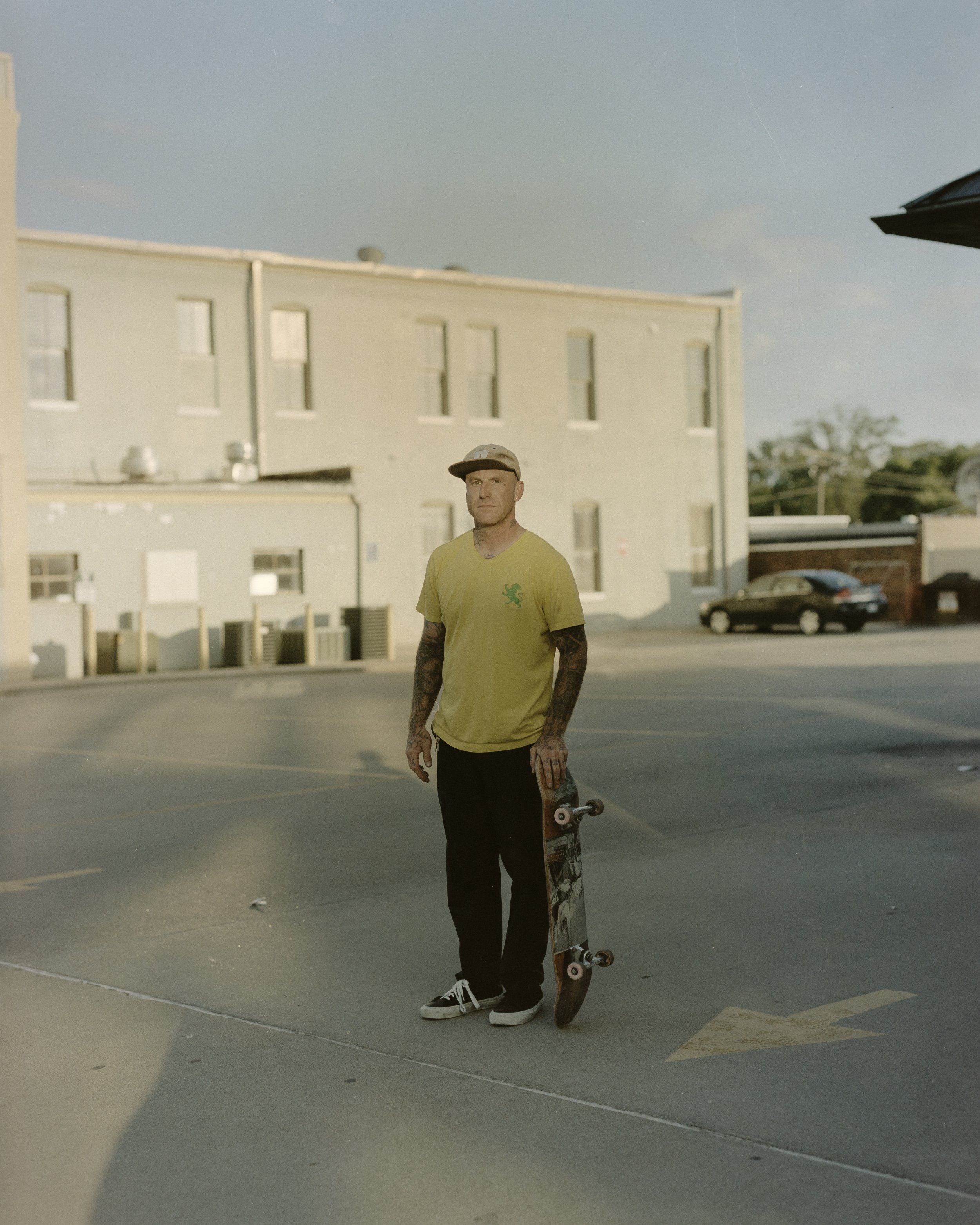 A man with tattoos, wearing a yellow T-shirt and a cap, holding a skateboard while standing in an empty parking lot.