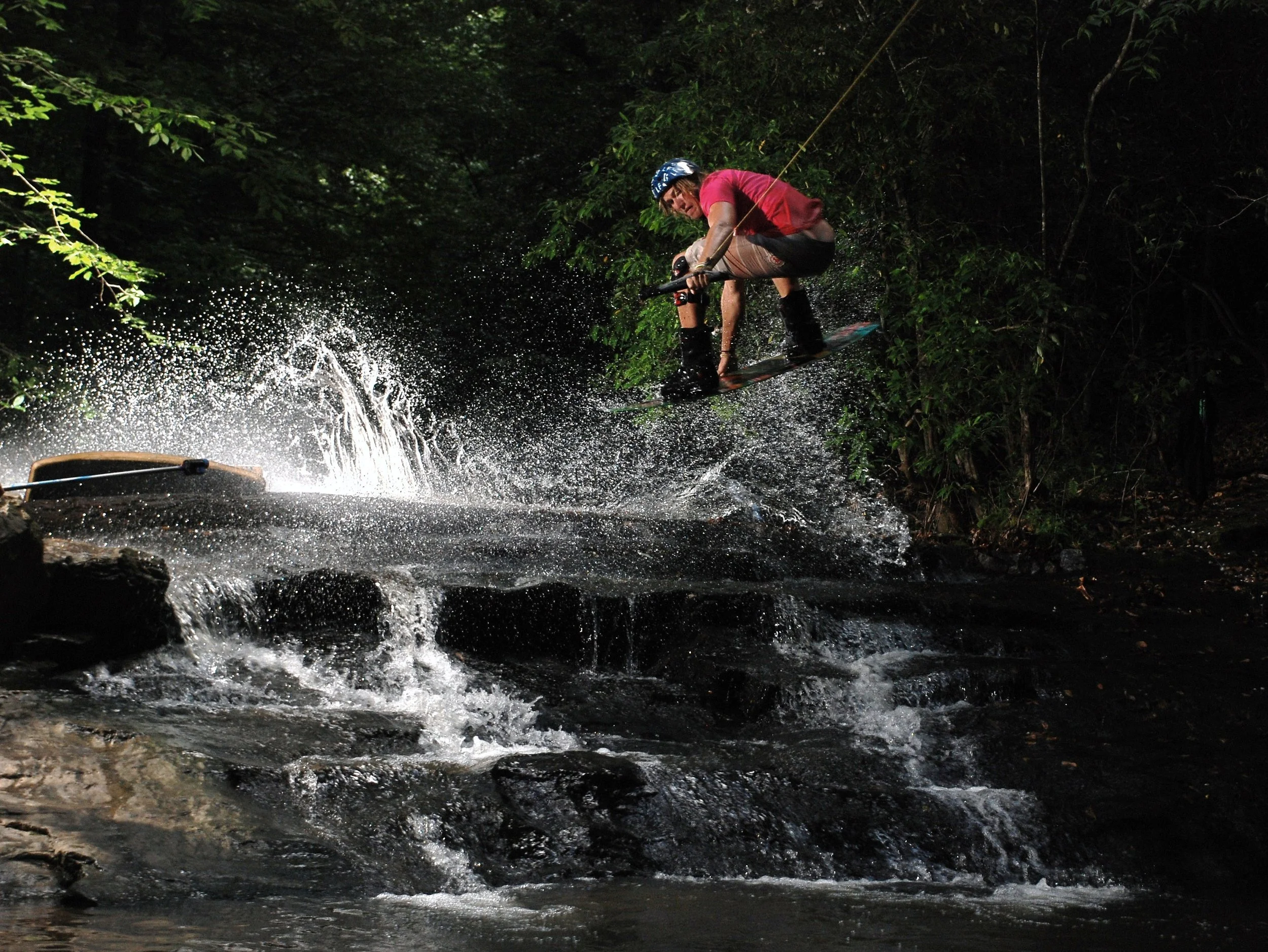 A person in a red shirt and helmet wakeskating over a small waterfall in a forested area at night.