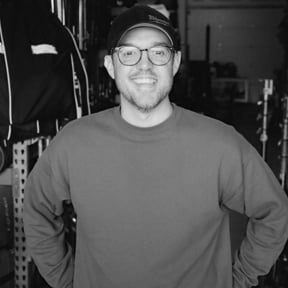 A smiling man wearing glasses, a cap, and a long-sleeve shirt, standing in a gym or fitness center.