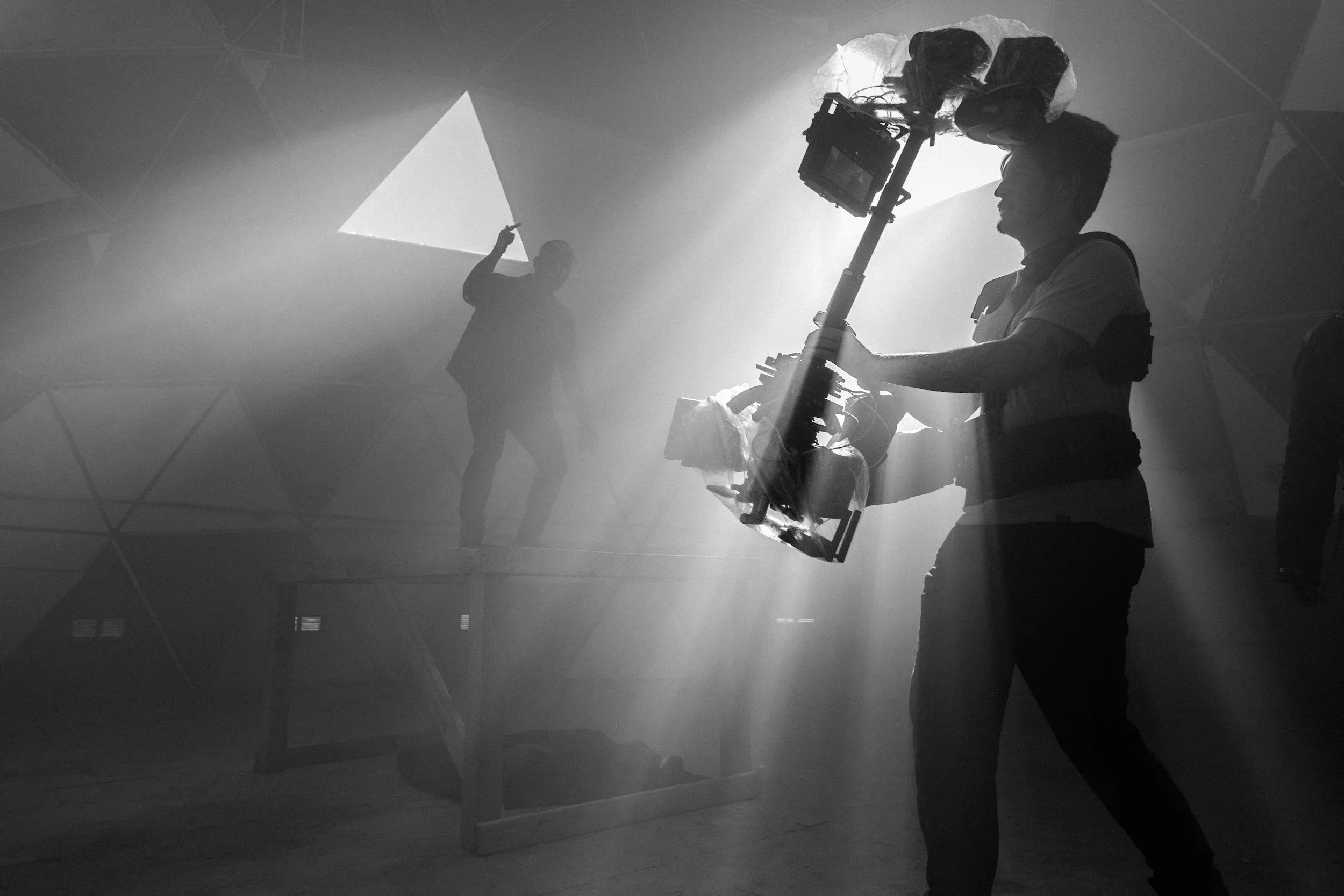 Black and white photo of a film crew filming a person on a raised platform inside a dome-shaped structure, with sunlight coming through a window.