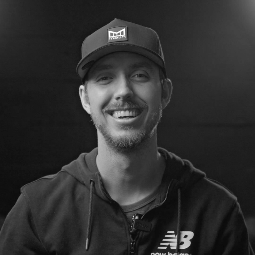 A young man smiling, wearing a black cap with a logo, a black zip-up hoodie with the New Balance logo, and a T-shirt underneath, in a black and white photo against a dark background.