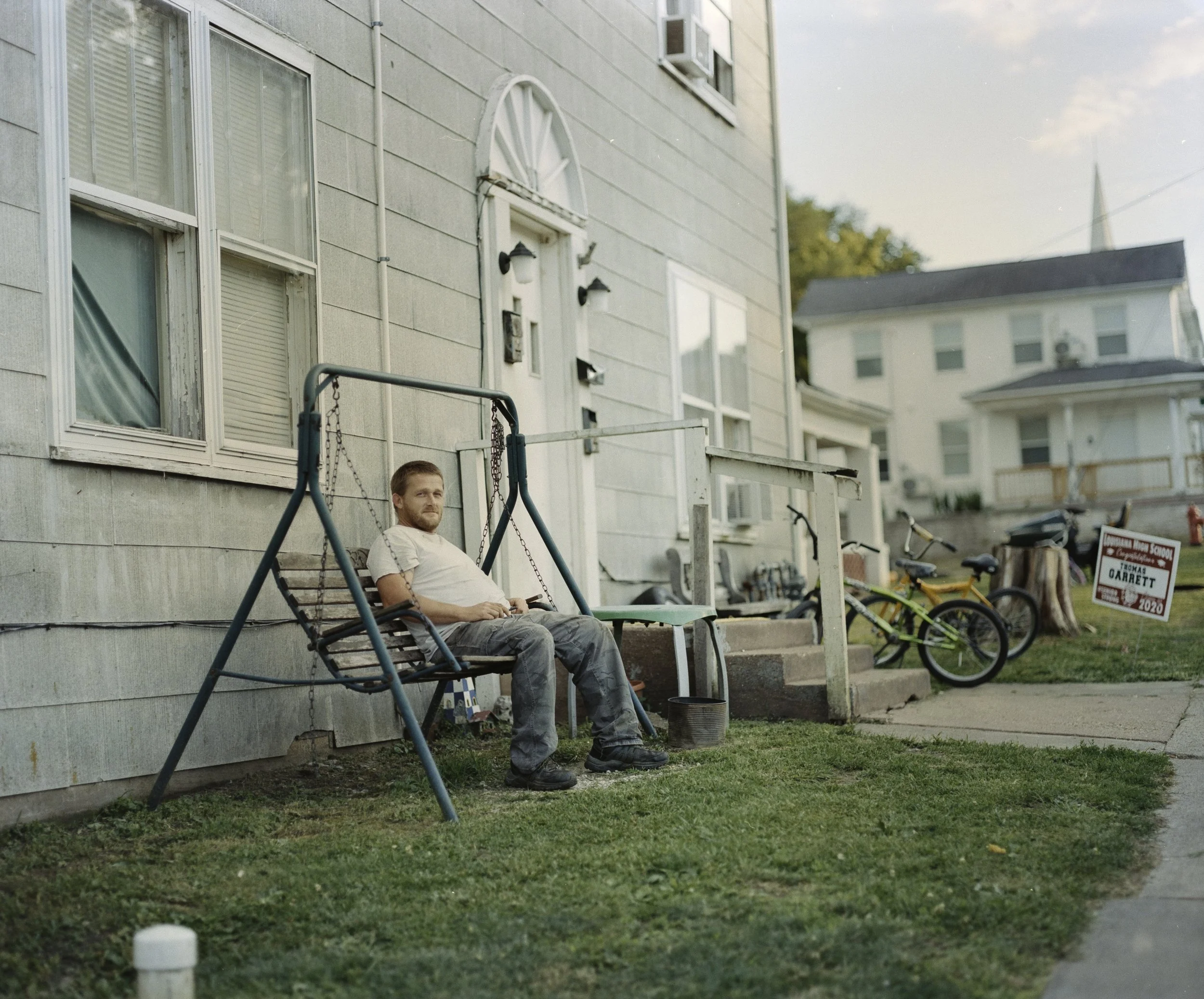 A man sitting on a swing in front of a house, with bicycles and a campaign sign nearby.