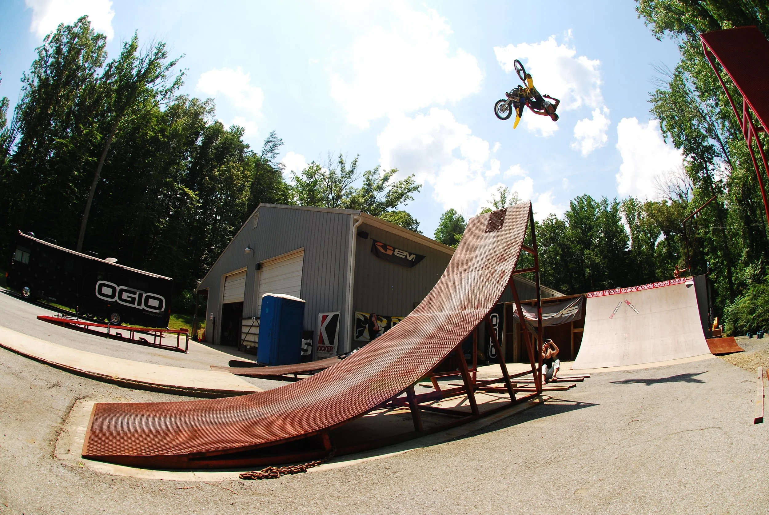 A person performing a stunt on a dirt bike mid-air at a skatepark with ramps, under a partly cloudy sky surrounded by trees.