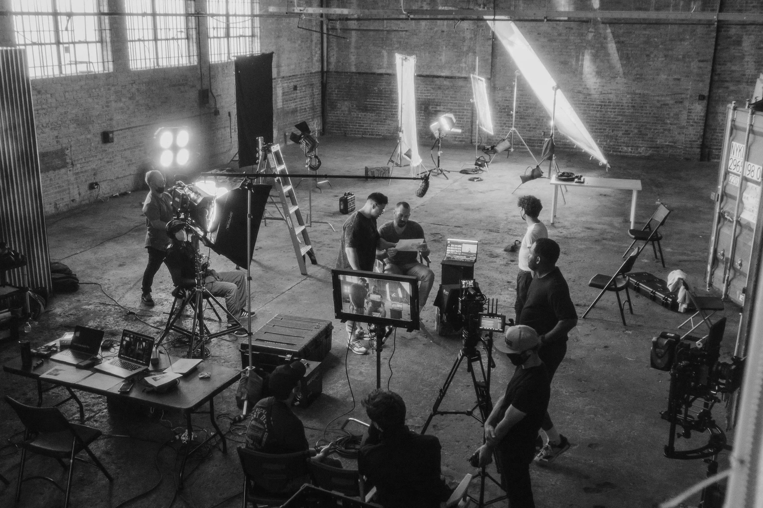 A film or video production set with crew members, cameras, lights, and equipment, in an industrial-style studio with brick walls and large windows.