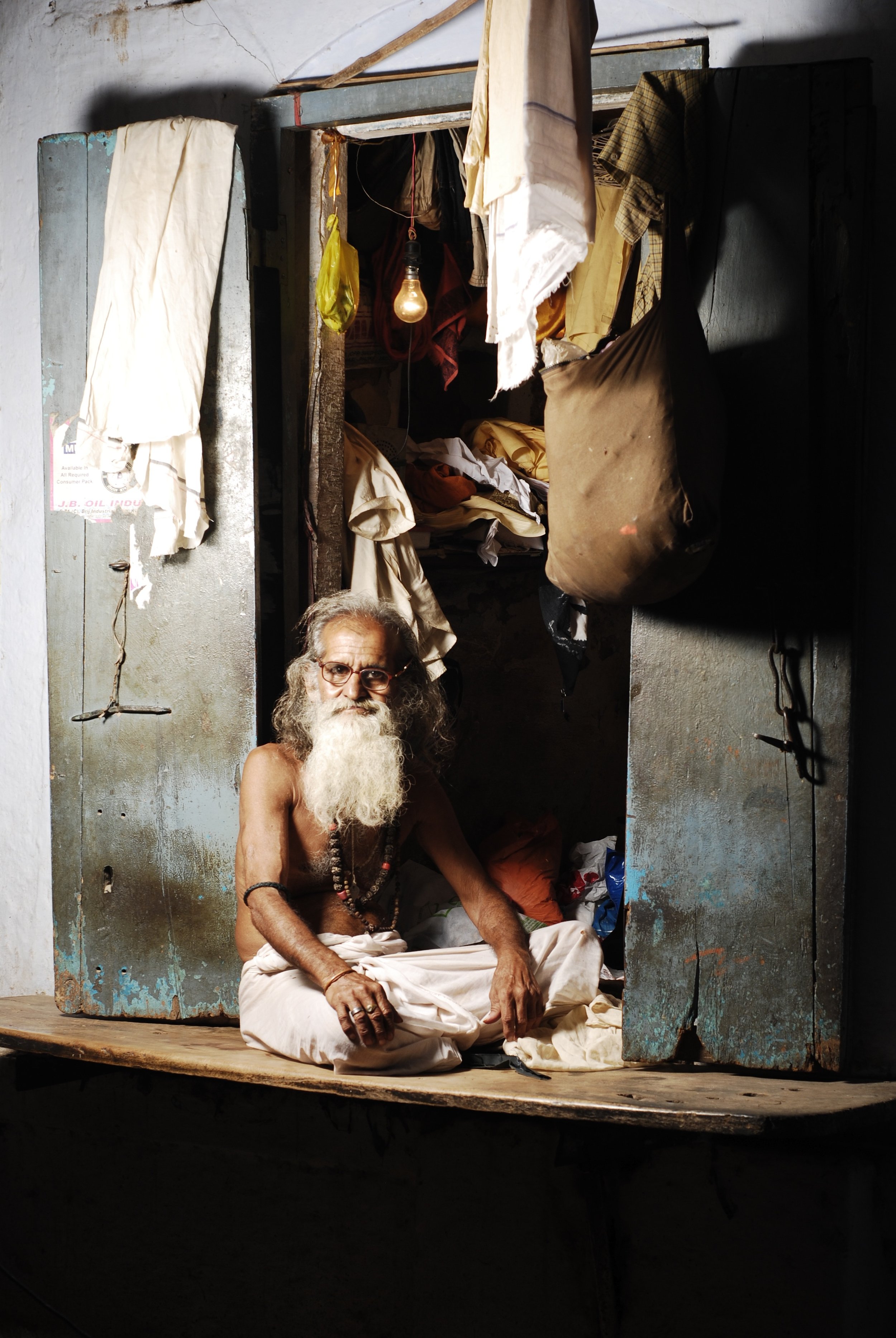 An elderly man with a long white beard and glasses sits cross-legged on a wooden platform inside a small, cluttered room. The room has a metal door and an open closet filled with clothes and other items. A hanging lightbulb illuminates the scene, casting shadows around.