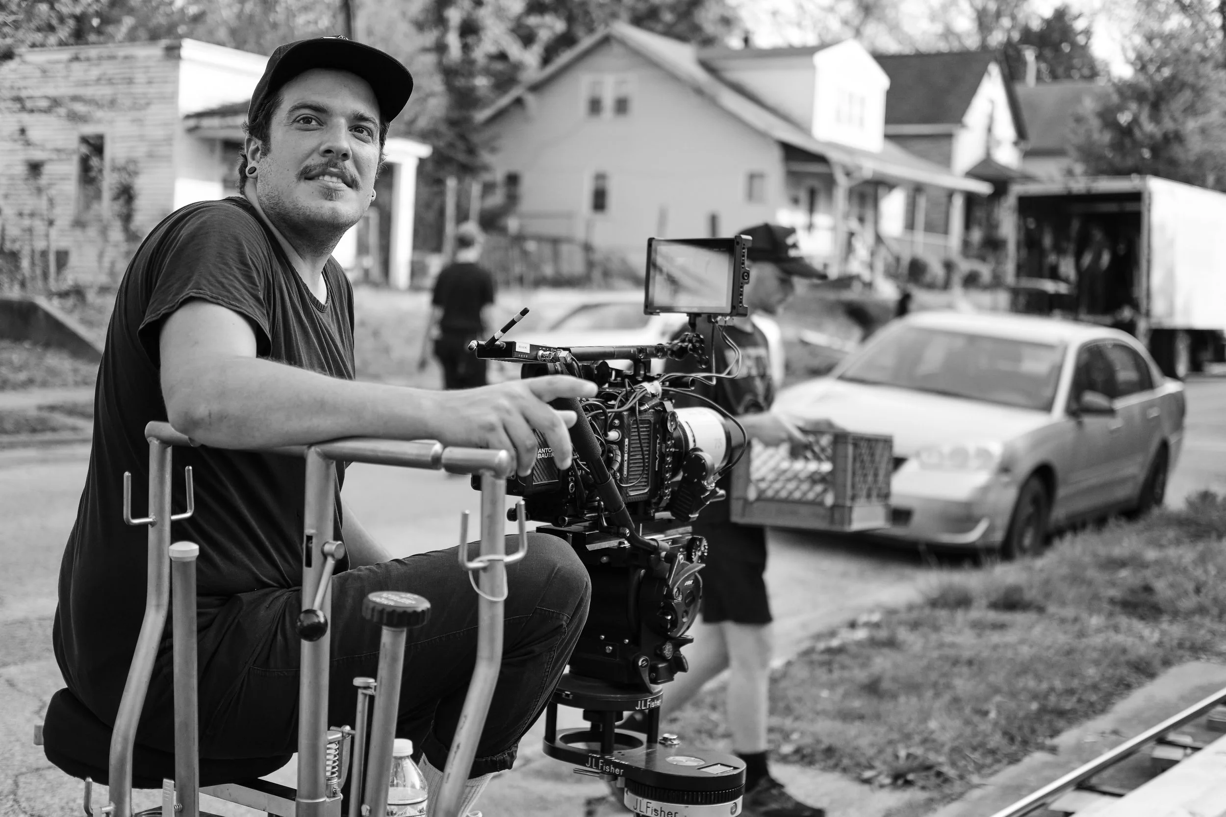 Black and white photo of a man sitting on a wheeled camera dolly, holding a professional film camera, outdoors on a sidewalk with houses and a car in the background.