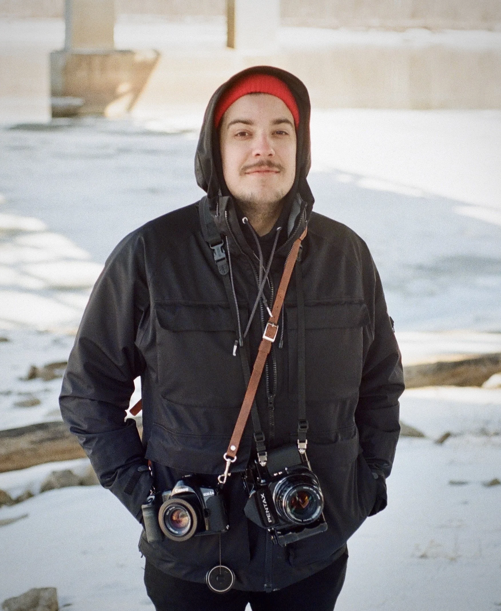 A young man in winter clothing, including a black jacket and red beanie under a hood, standing outdoors on snow with a camera hanging around his neck.