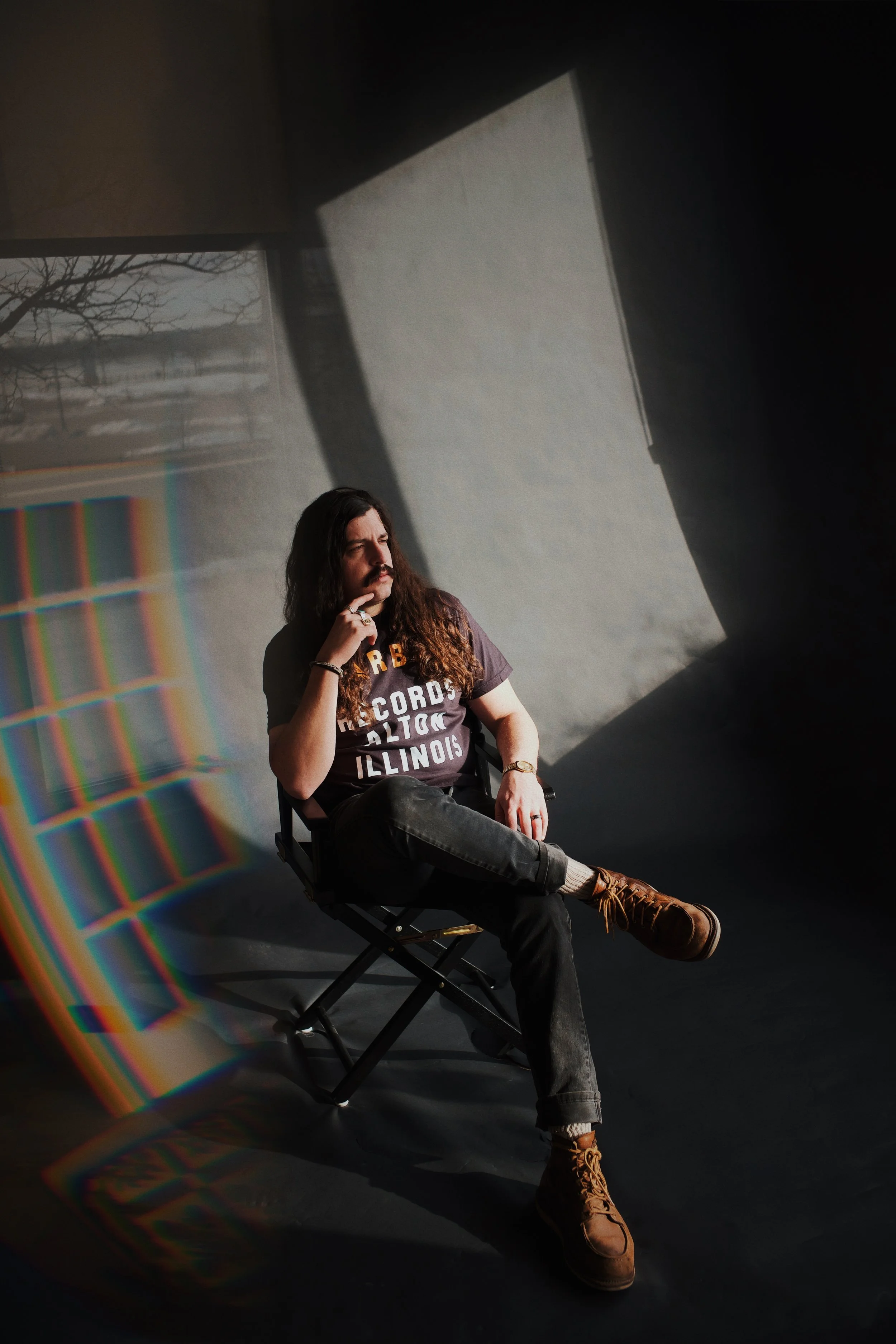 A man with long hair and a beard, wearing a dark T-shirt with words printed on it, black pants, and brown boots, sitting in a black folding chair in front of a wall with sunlight casting geometric shadows. Light also creates a rainbow reflection on the floor nearby.