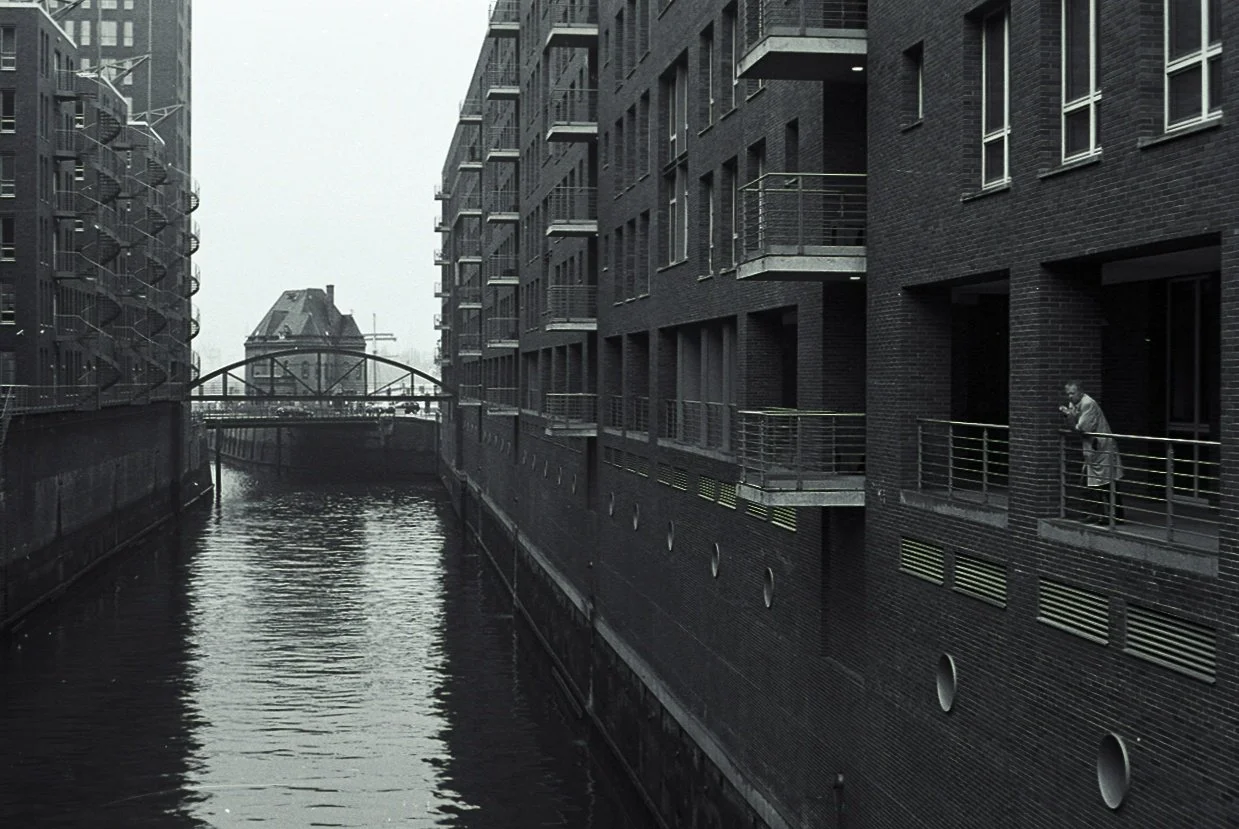 A canal with dark brick apartment buildings on both sides and a small bridge in the distance, with a person standing on a balcony looking at a phone.