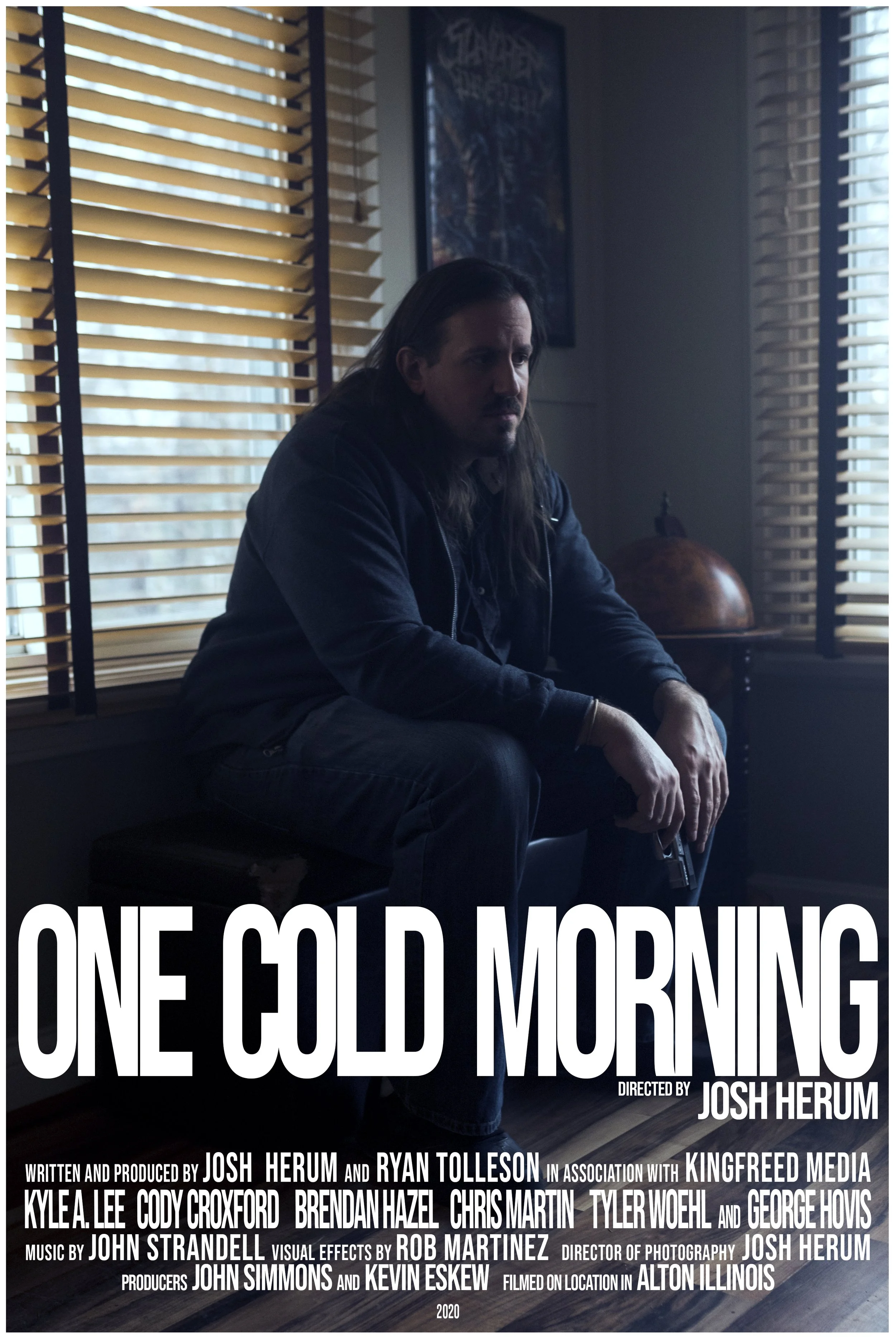 A man with long hair, dressed in dark clothing, sitting on a bench or seat in a dimly lit room with wooden blinds and a globe on a table behind him. A poster is on the wall behind him. The image is a promotional poster for a film titled 'One Cold Morning,' directed by Josh Herum.