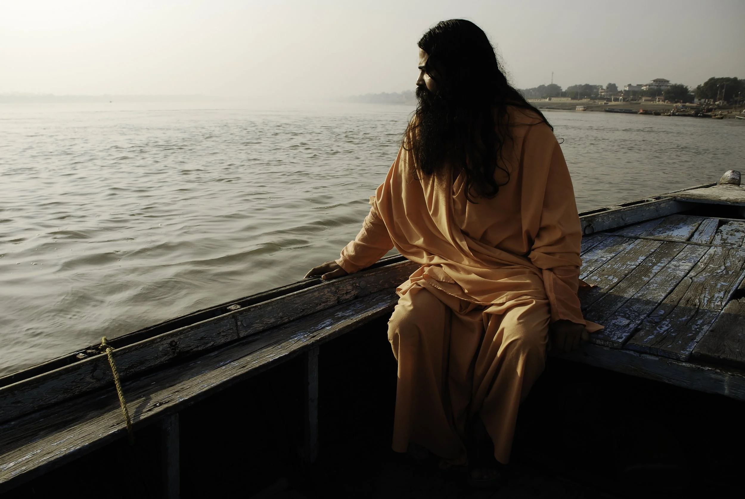 A man dressed in orange robes sitting on a wooden boat, looking out over a calm river during sunset or sunrise.