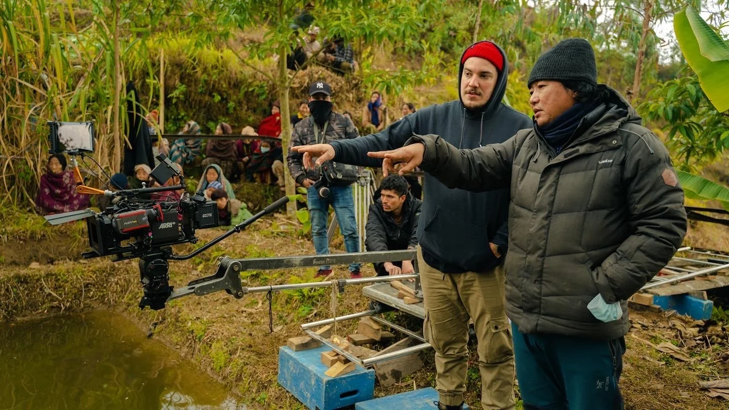 Filmmakers on a set in a tropical outdoor location, working with a camera on a steady rig, while an audience or crew members watch in the background.