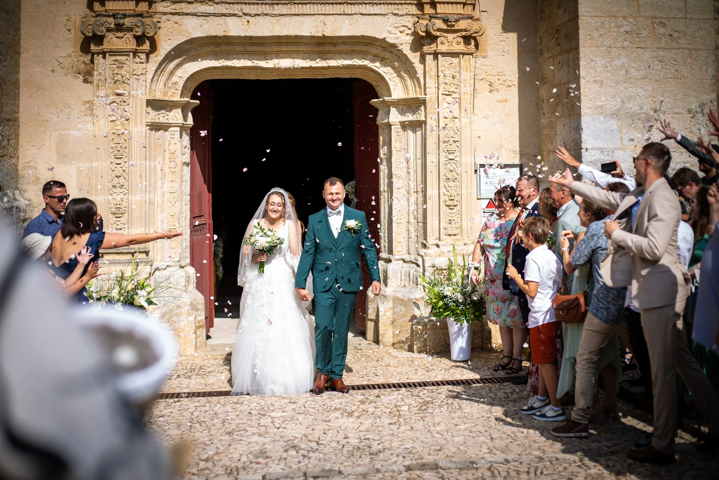 Un couple de mariés sortant d'une église en pierre, entourés de leurs invités qui leur lancent des pétales de fleurs.