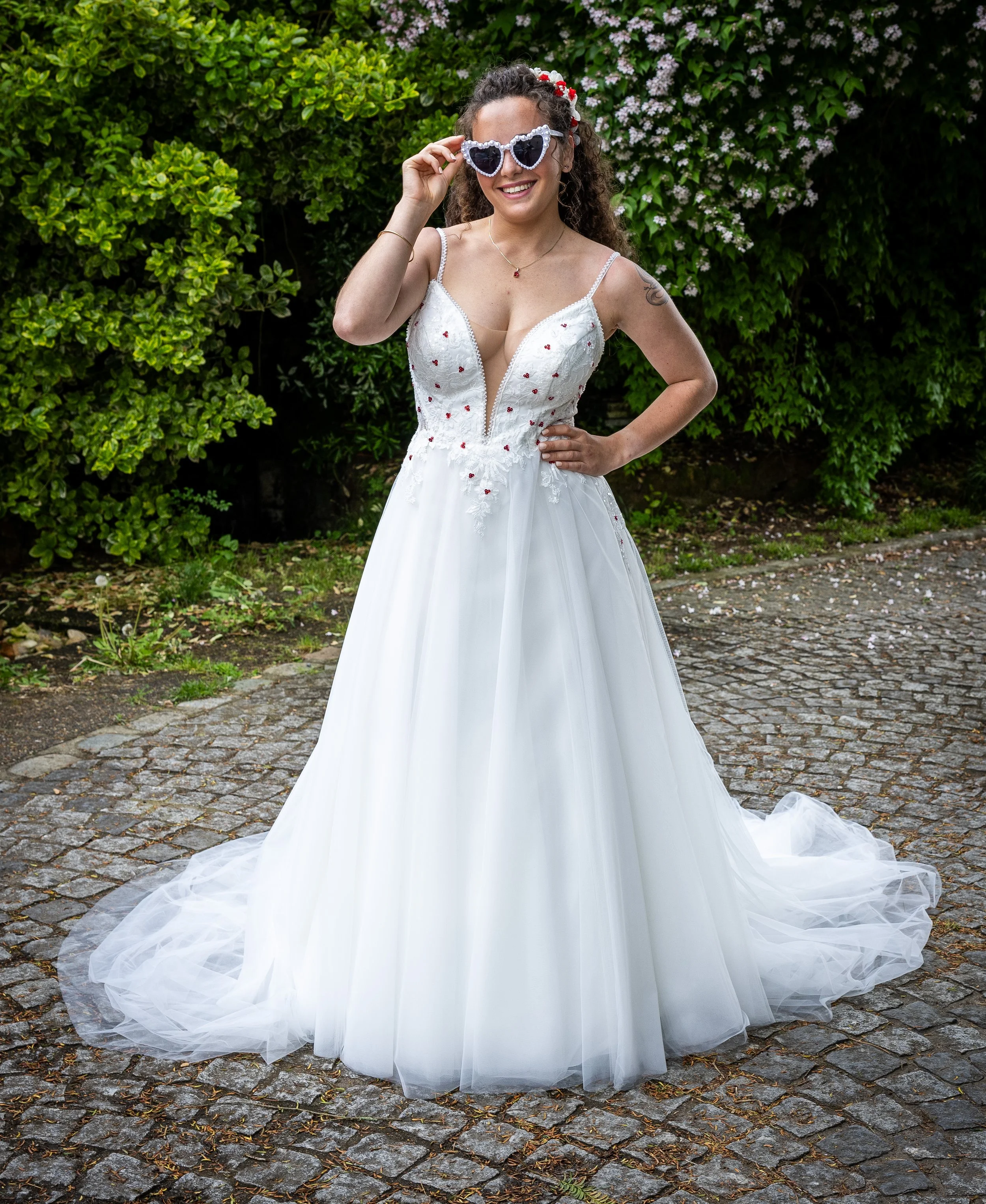 Une femme en robe de mariée blanche avec des détails rouges, portant des lunettes de soleil en forme de cœur, souriant, devant un buisson vert avec des fleurs roses et une allée pavée.