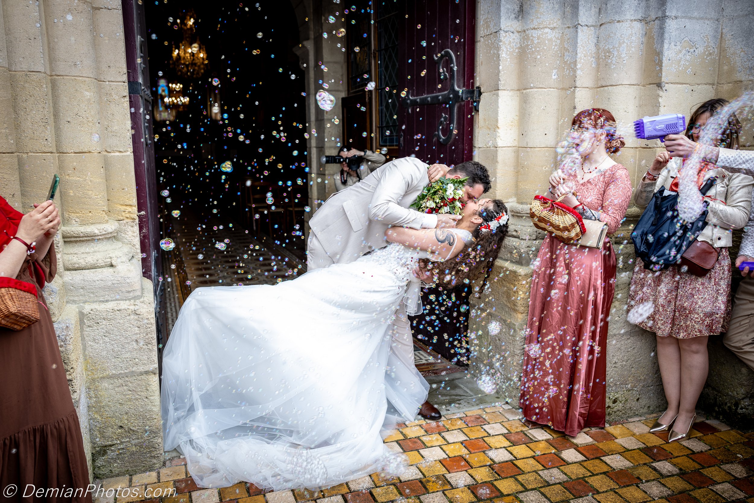 Un couple de mariés s'embrasse devant l'entrée d'une église, entourés de personnes qui les prennent en photo et lancent des bulles de savon, célébrant leur mariage.