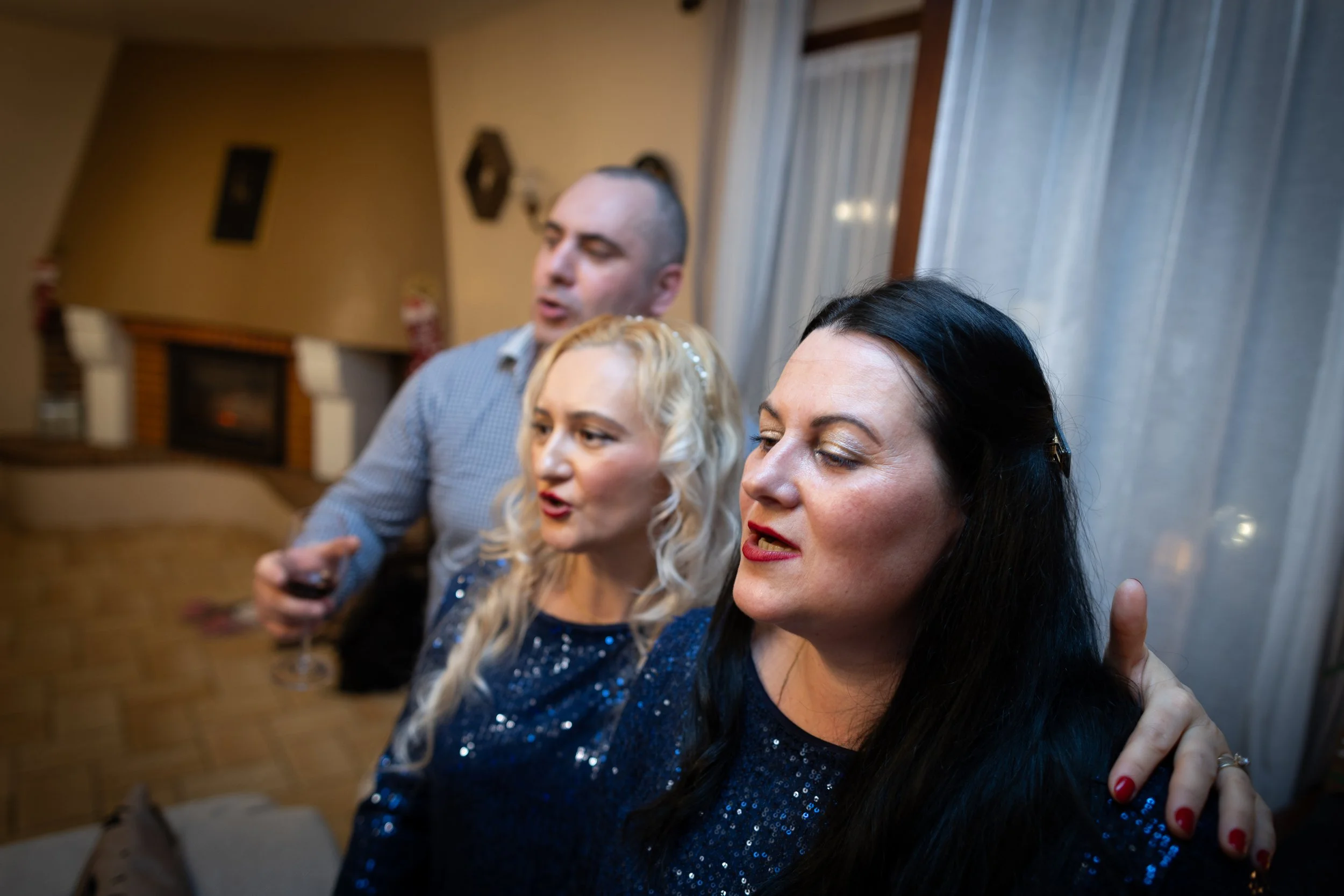 Trois personnes, deux femmes et un homme, qui semblent chanter lors d'une réunion sociale. Les femmes sont en premier plan, une porte de fenêtre avec des rideaux en arrière-plan.