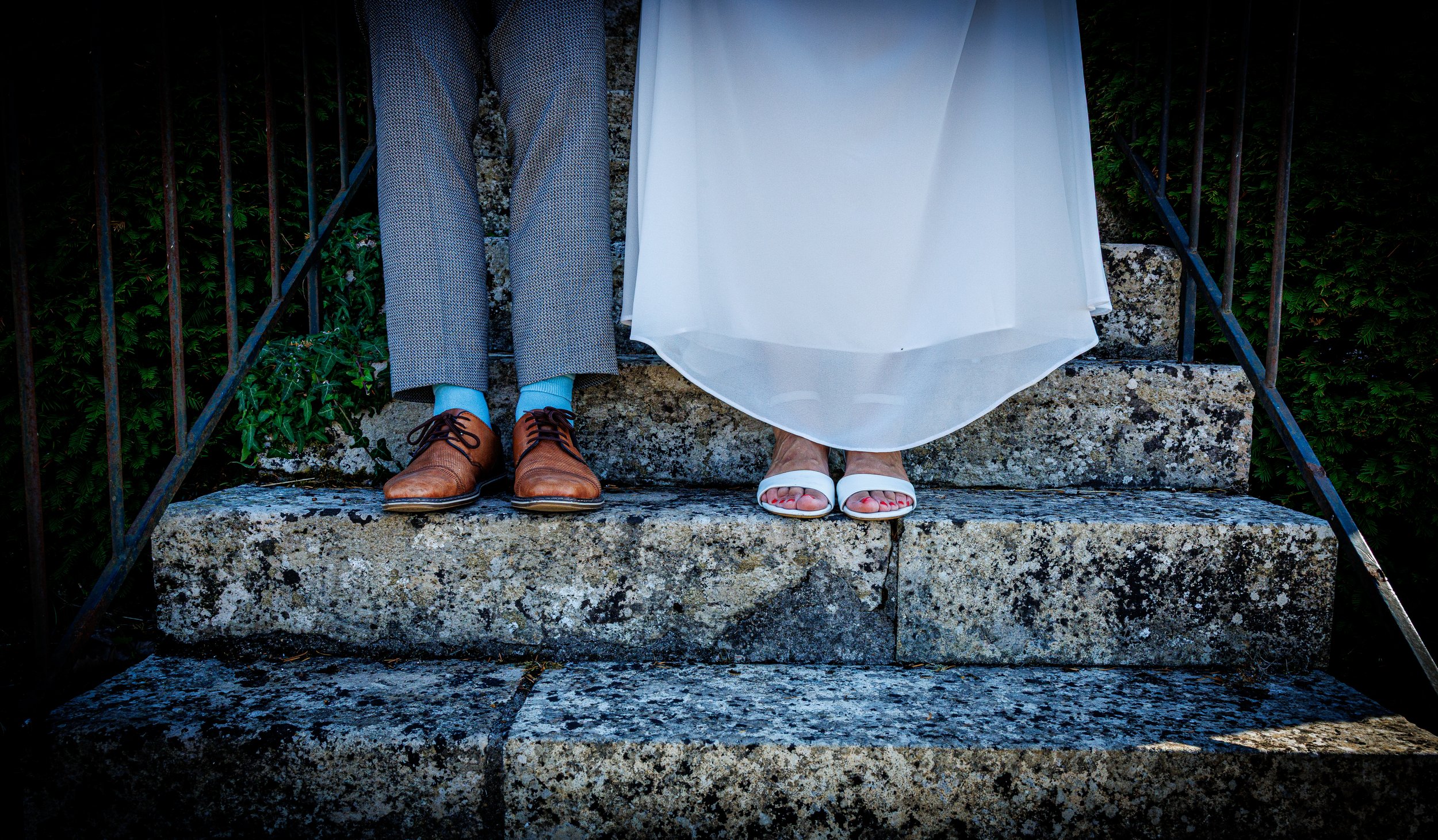 Les jambes et les chaussures de deux personnes assises sur une marche en pierre, dont une personne porte des chaussures en cuir marron et des chaussettes bleues, et l'autre porte des sandales blanches et une robe blanche. La photo ne montre pas leur tête ni leur visage, et il y a des plantes à l'arrière-plan.