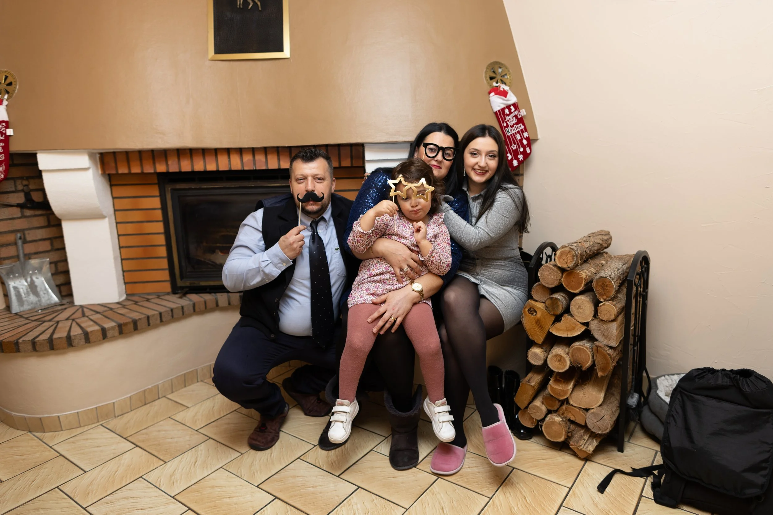 Groupe de quatre personnes posant devant une cheminée décorée de chaussettes de Noël, avec un panier de bois de chauffage à côté, dans une pièce chaleureuse.