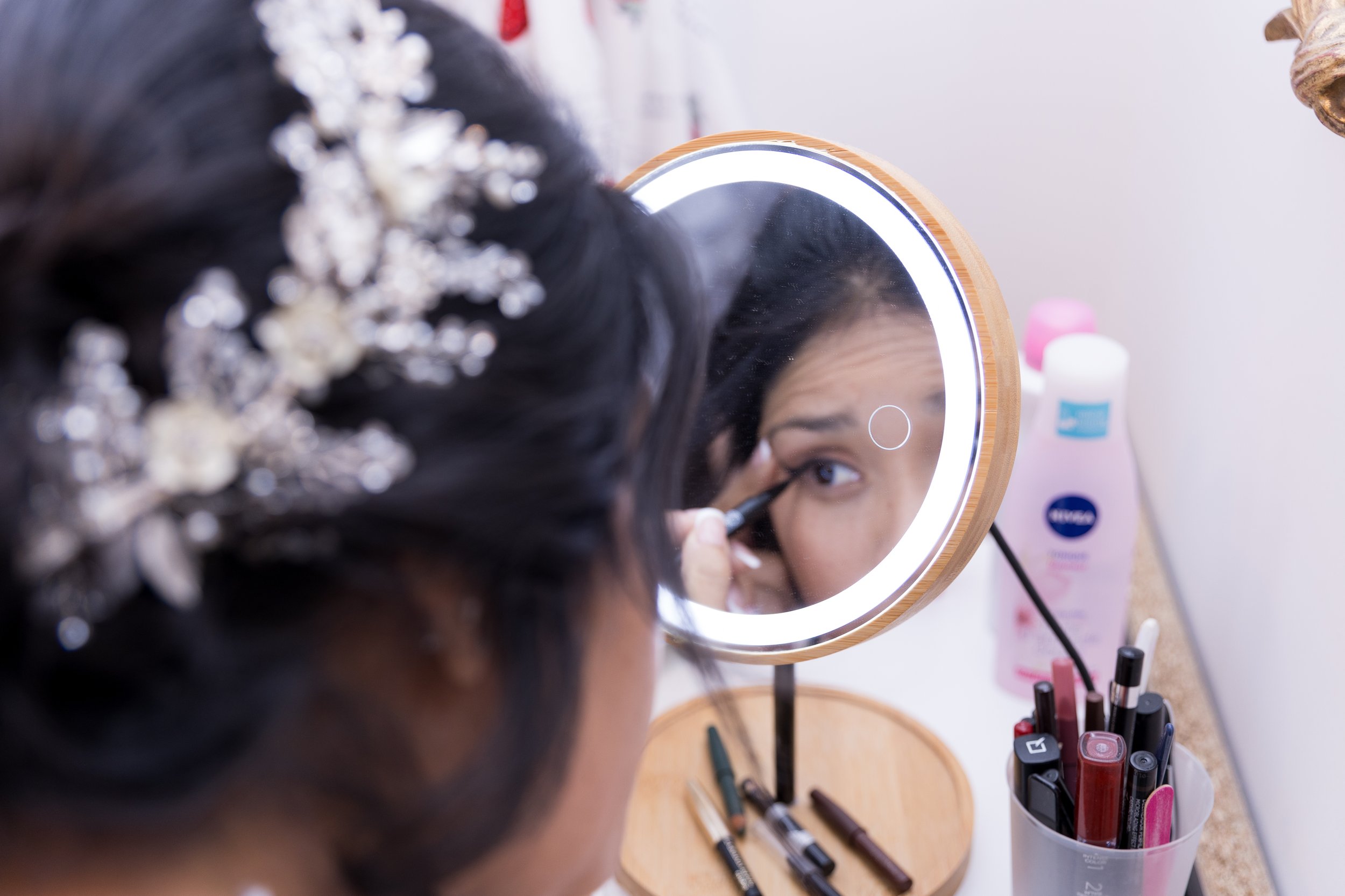 Une femme se maquillant dans un miroir rond avec un cadre en bois, entourée de divers produits cosmétiques sur une table.