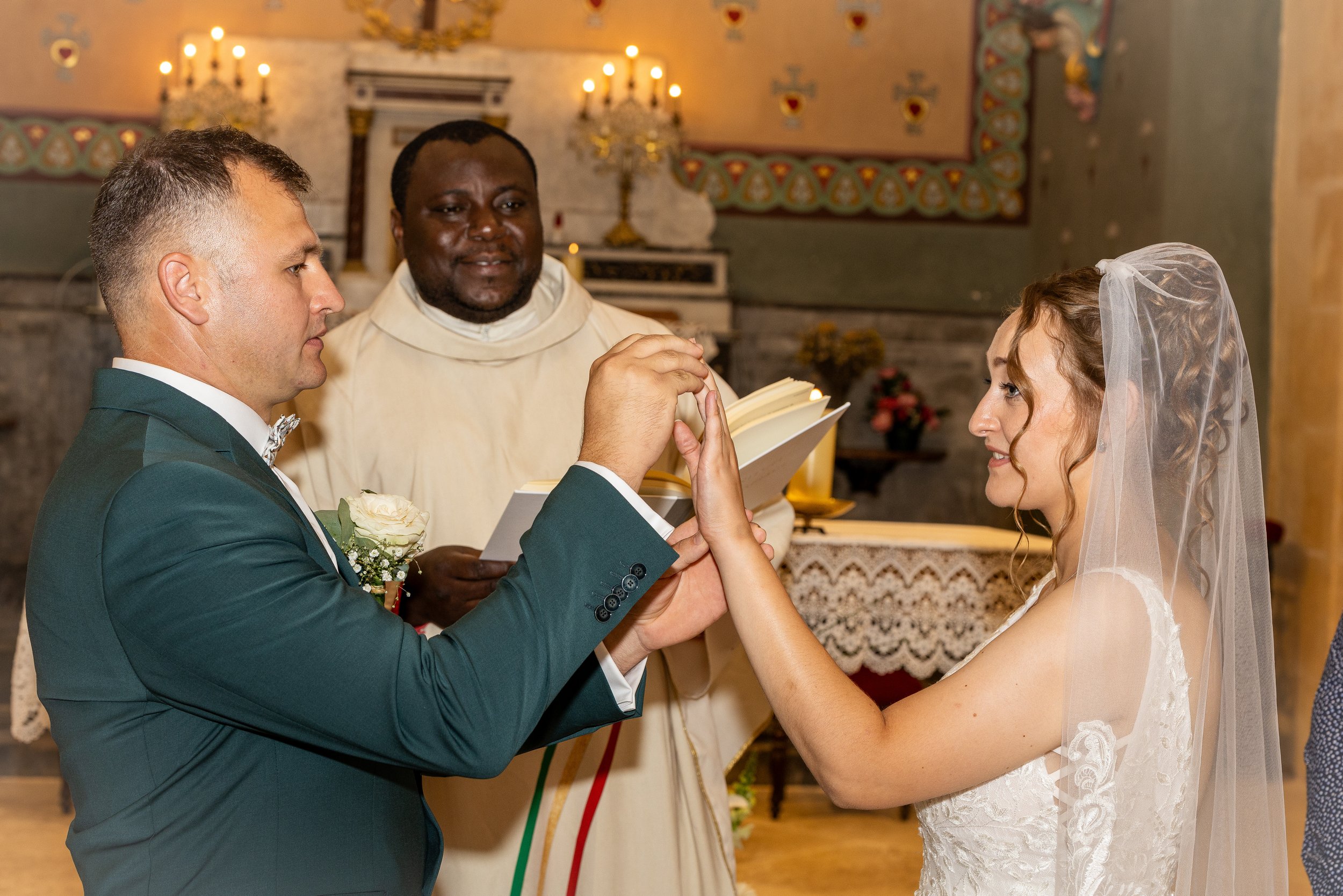 Un couple de mariés échangeant leurs vœux lors de la cérémonie religieuse, avec un prêtre en arrière-plan dans une église ornée.