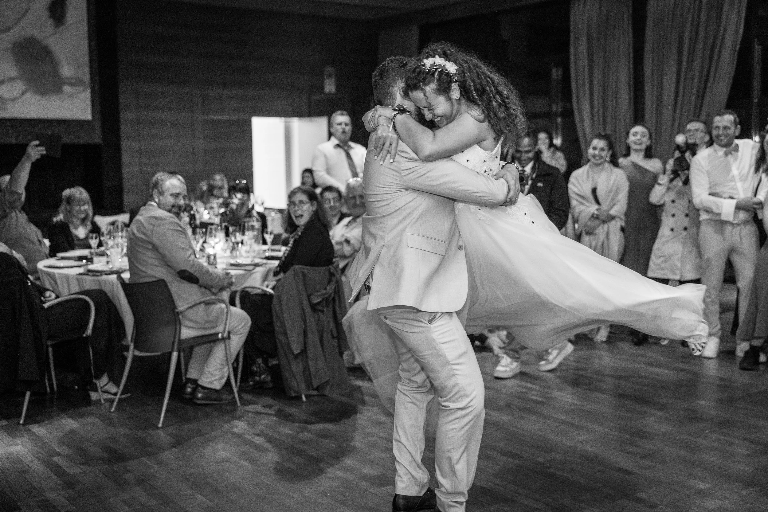 Un couple danse lors d'une réception, entourés d'invités assis à des tables et applaudissant. La scène est en noir et blanc.