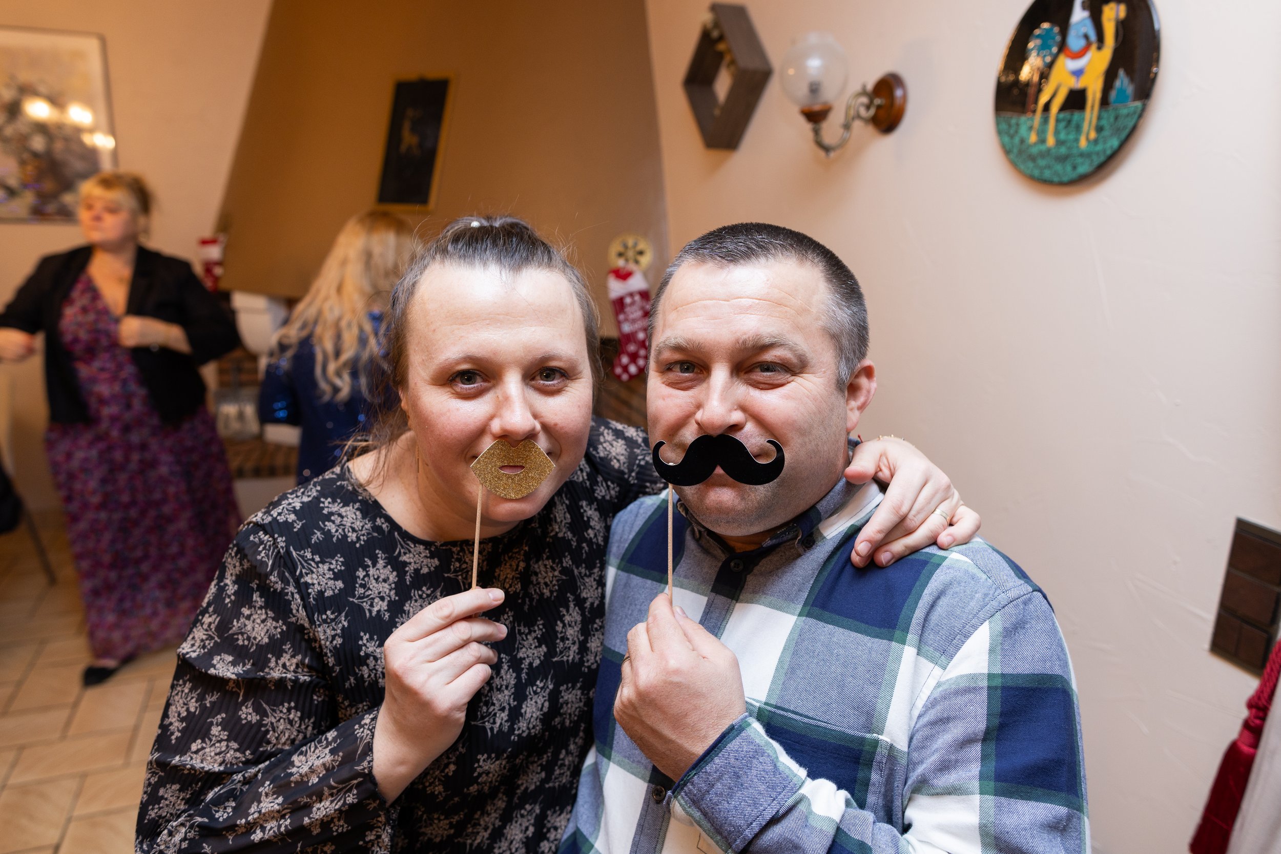 Deux personnes, un homme et une femme, posent avec des accessoires de fête pour une photo, dans une ambiance festive. En arrière-plan, d'autres invités assistent à l'événement.