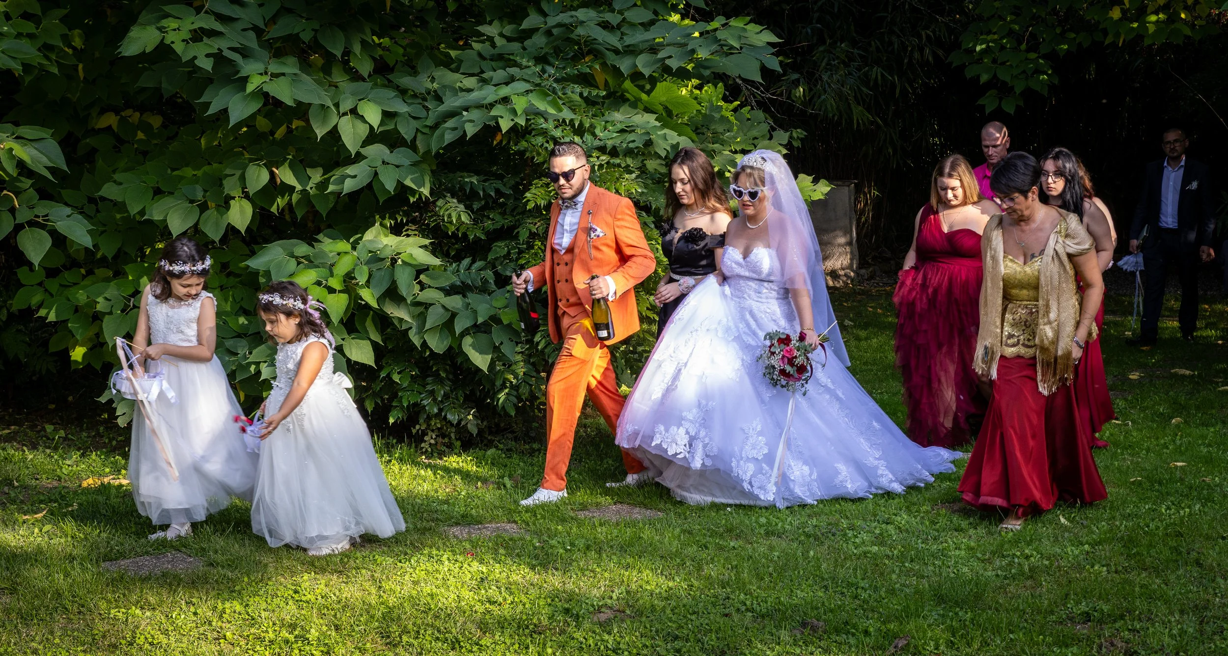 Un mariage se déroule devant une église en pierre. La mariée en robe blanche et le marié en costume orange saluent la foule. Des invités applaudissent et sourient.