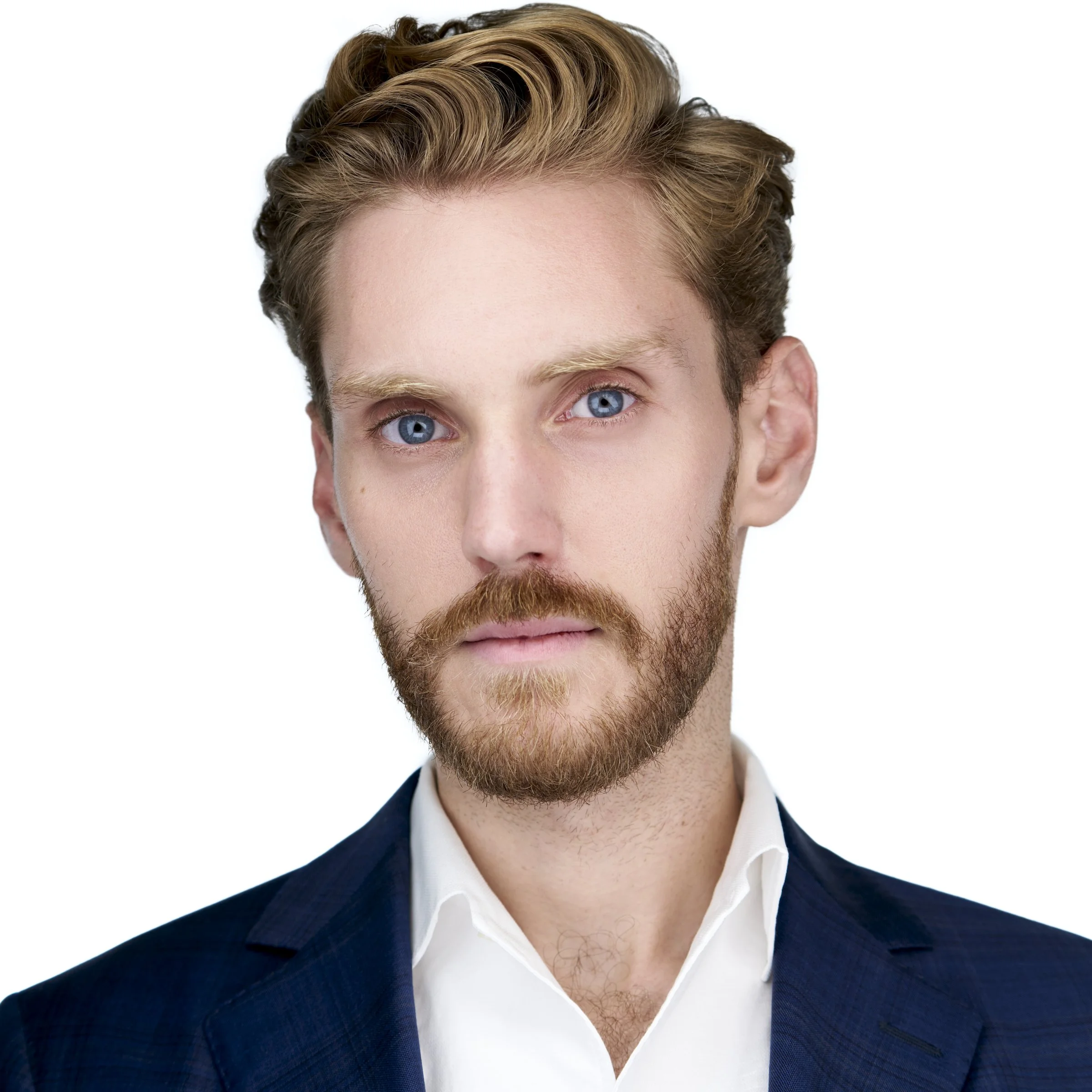 Portrait of a young man with blue eyes, reddish-brown hair, a beard, wearing a navy blue suit and a white dress shirt against a white background.