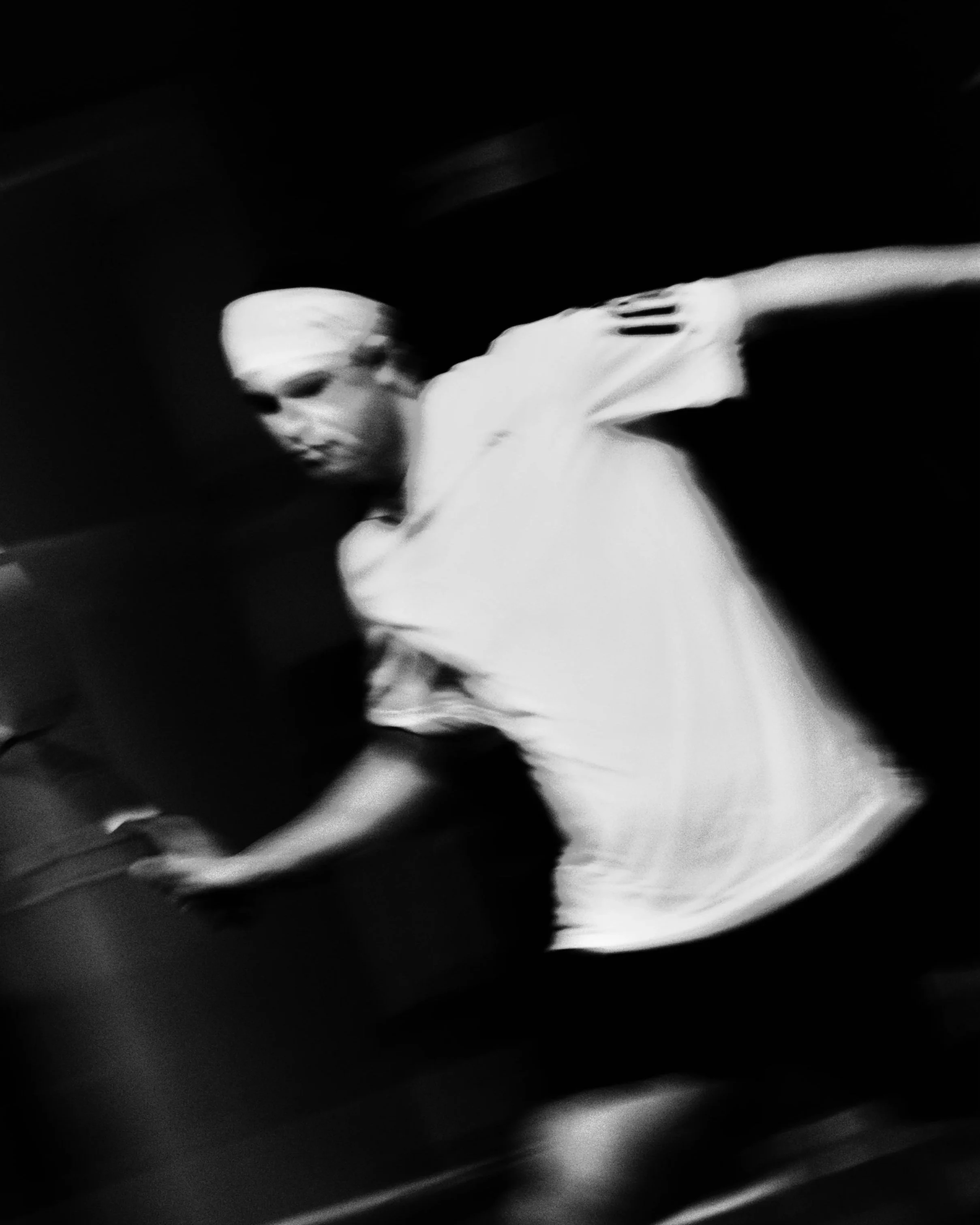 Black and white photo of a woman in motion, wearing a white t-shirt, with short blond hair, in a dark environment, possibly walking or reaching for something.