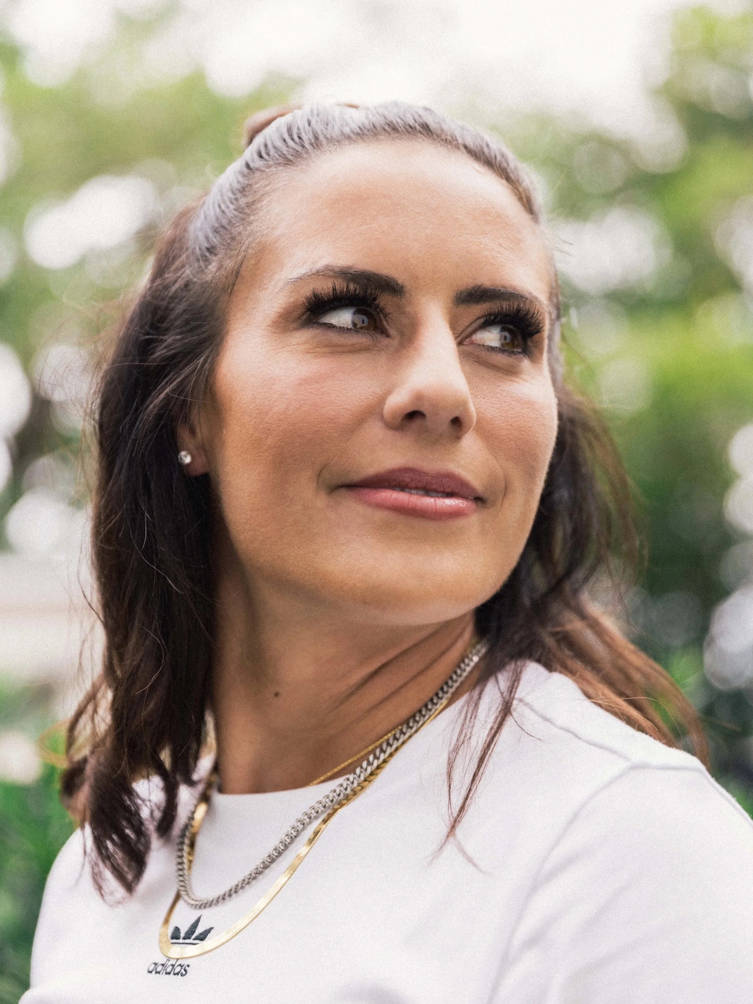 Close-up of a woman with dark hair tied back, wearing jewelry and a white shirt, outdoors with blurred greenery in the background.