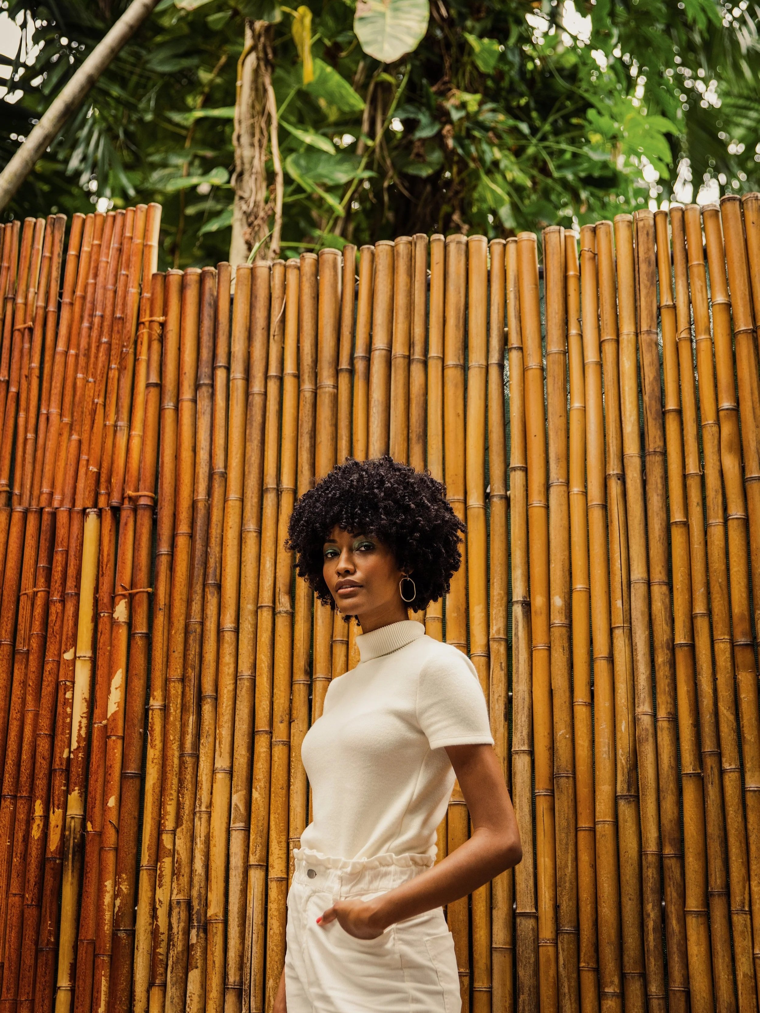 A woman with curly hair wearing a cream-colored turtleneck and white pants standing in front of a bamboo fence with tropical greenery in the background.