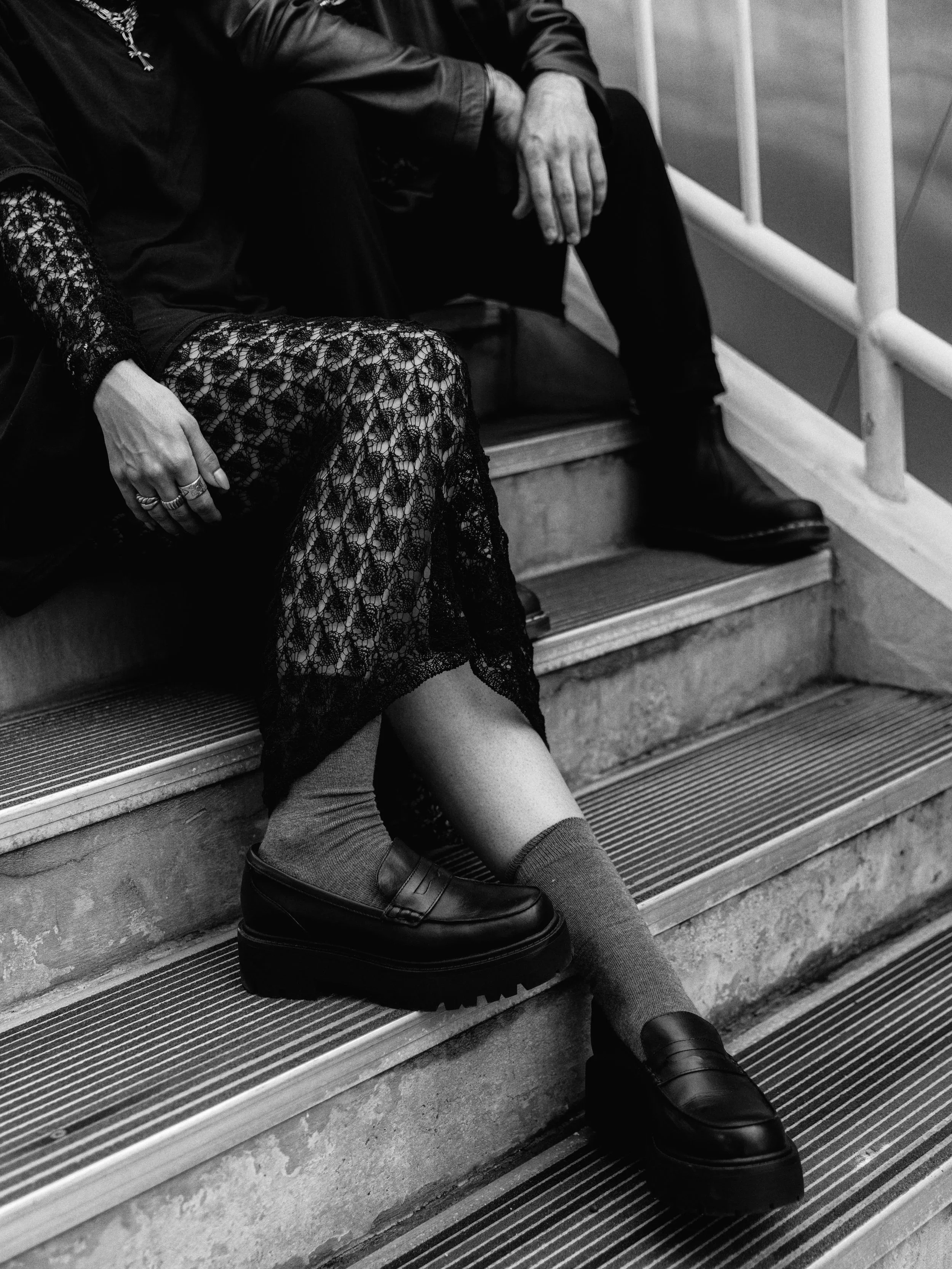 Close-up of two people sitting on stairs, with focus on one person's legs and shoes, wearing platform loafers and socks, and the other person's hand on the lap. The image is in black and white.