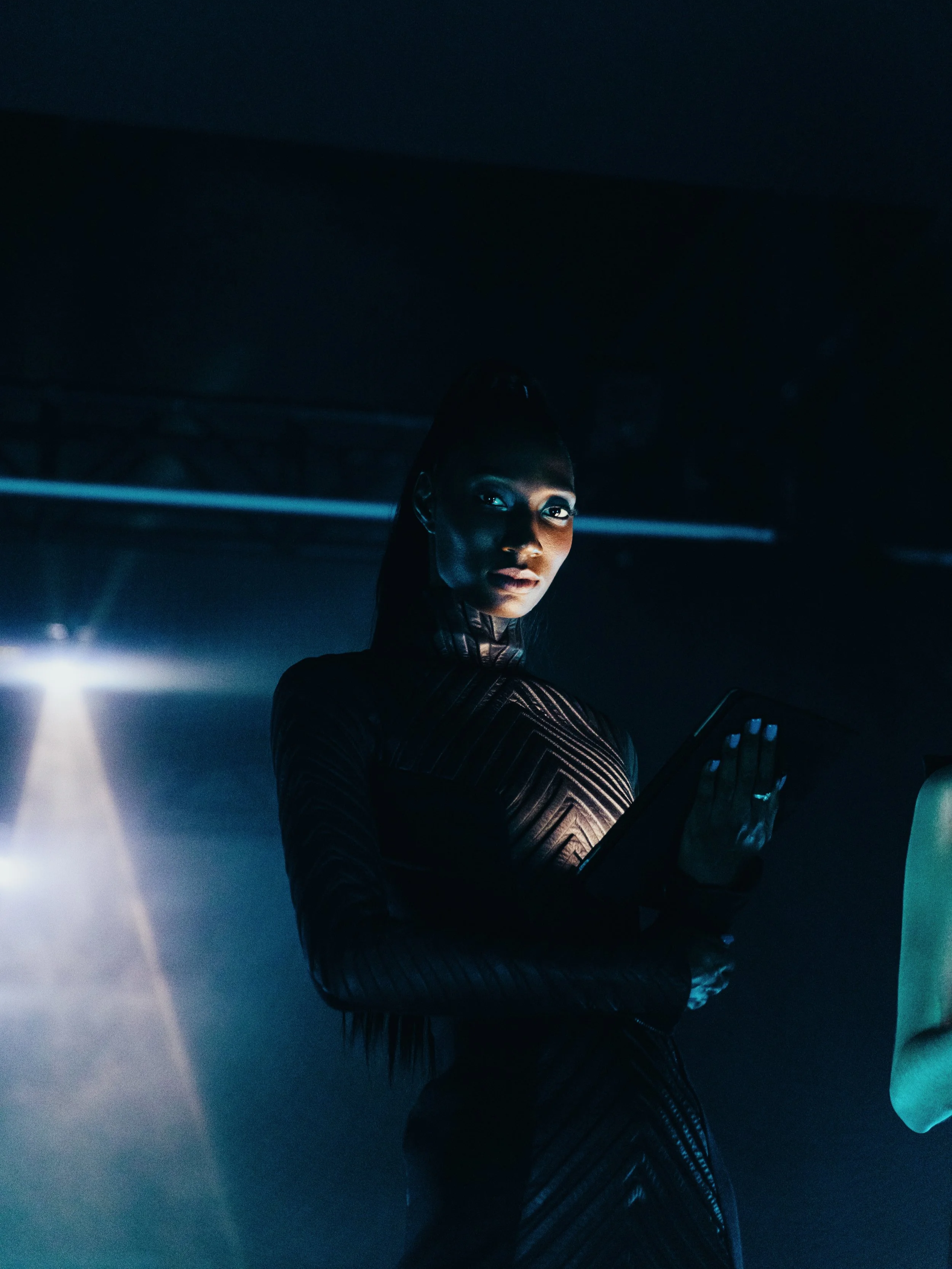 A woman in a dark, textured dress holding a tablet in a dimly lit environment with a blue hue and faint light beams.