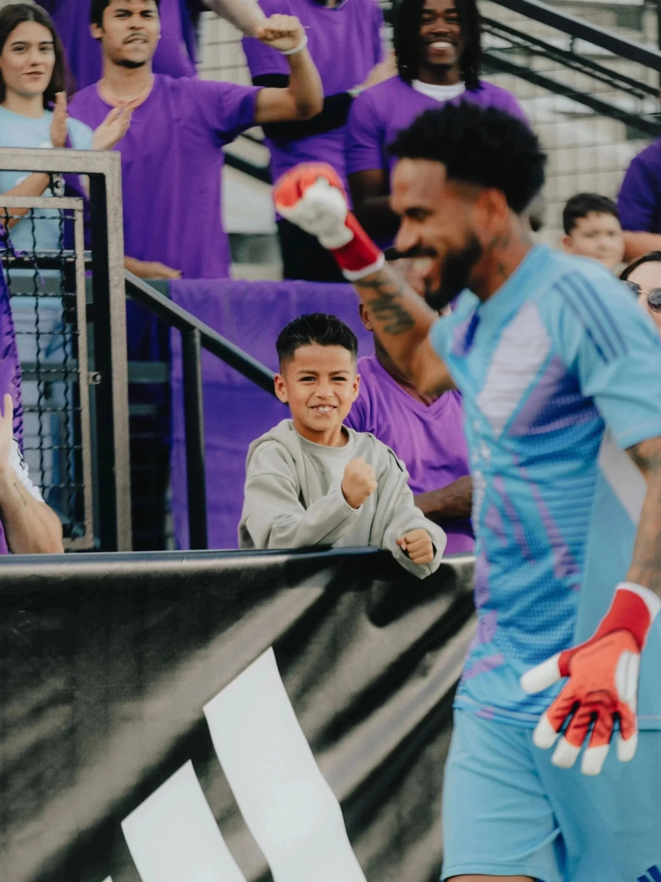 A young boy stands behind a black barrier, smiling and making a fist gesture. A soccer player in a light blue jersey and orange gloves is in the foreground, smiling and gesturing with a fist. Several people wearing purple shirts are in the background