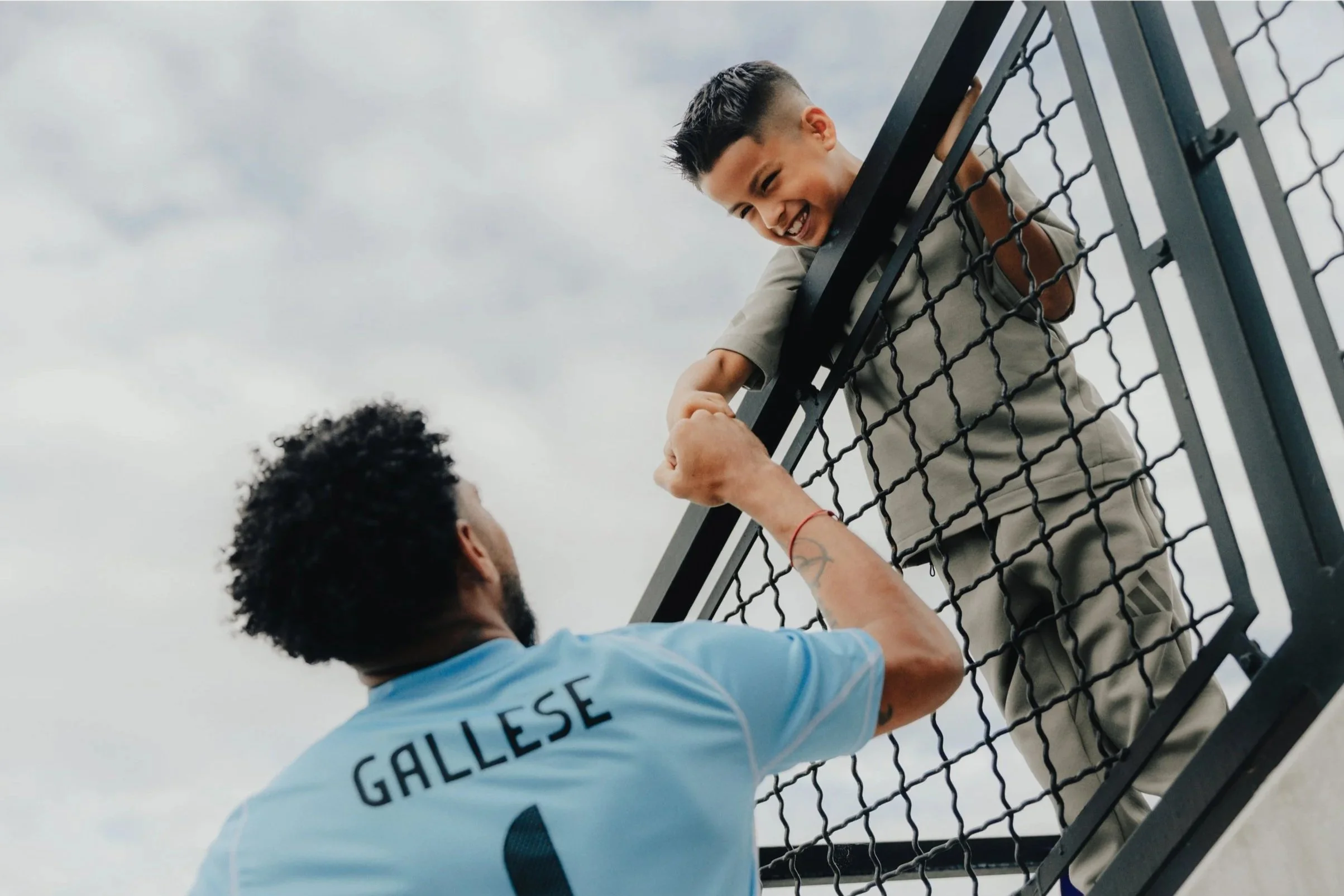 A young boy on a soccer field leaning over a fence, smiling and holding hands with a man wearing a light blue soccer jersey with the name "Gallese" on the back.