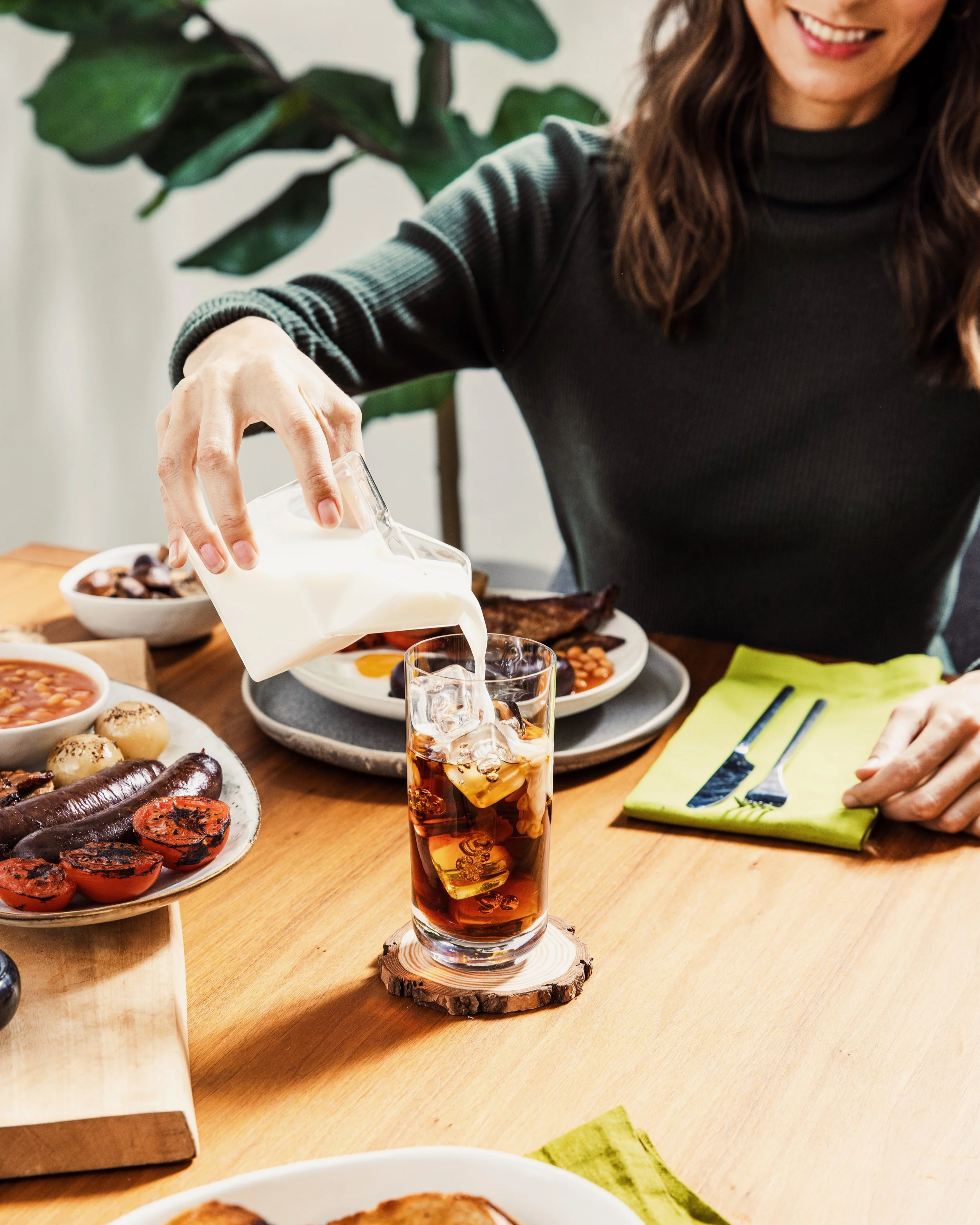 A woman in a long-sleeved black top pours milk into a glass of iced tea on a wooden table with various breakfast foods.
