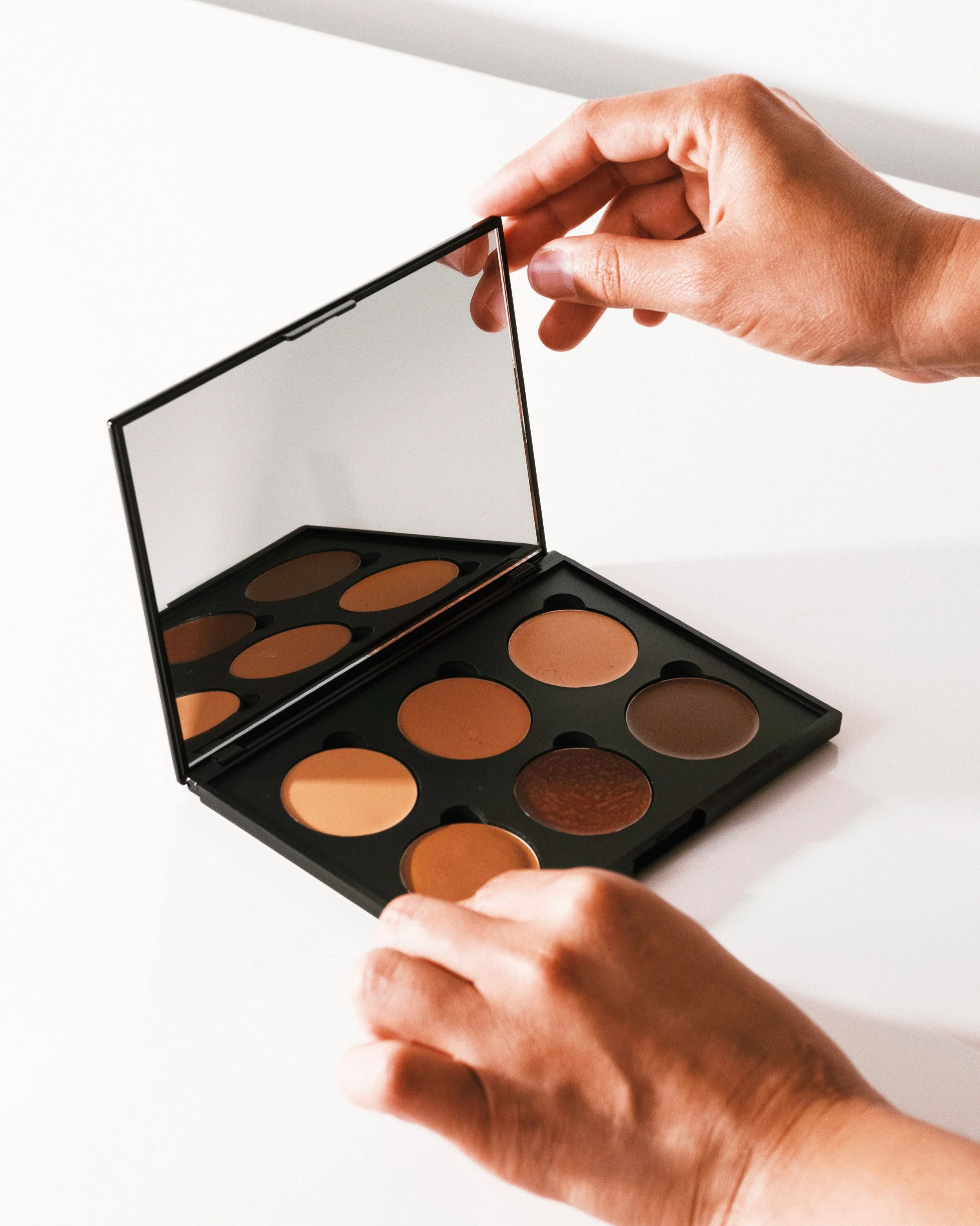 Open makeup palette with eyeshadow shades and a mirror, held by hands against a white background.