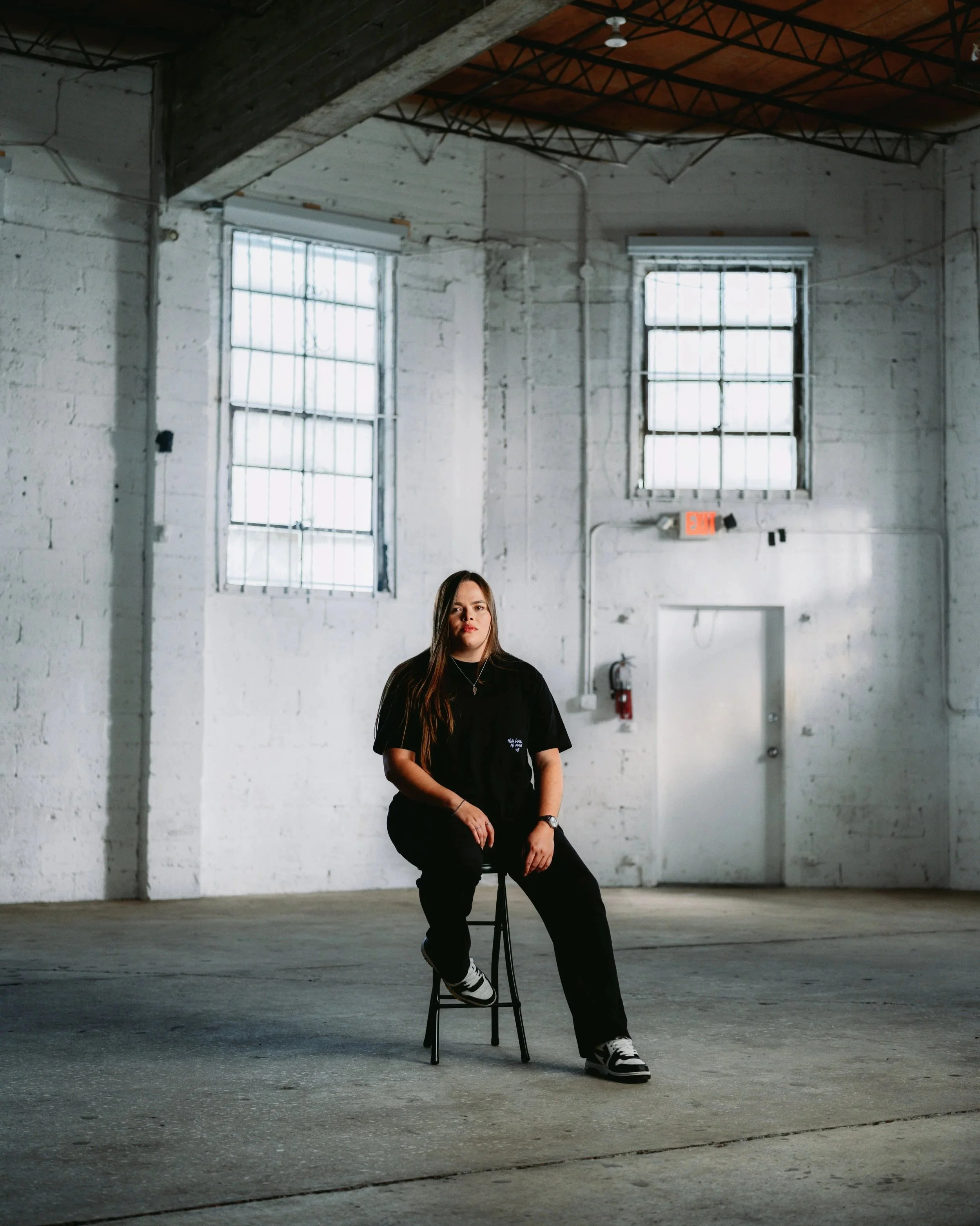 A young woman with long brown hair is sitting on a tall black stool in an industrial-style room with white brick walls, large windows, and exposed wooden beams on the ceiling. She is wearing a black t-shirt, black pants, and sneakers, and is looking 