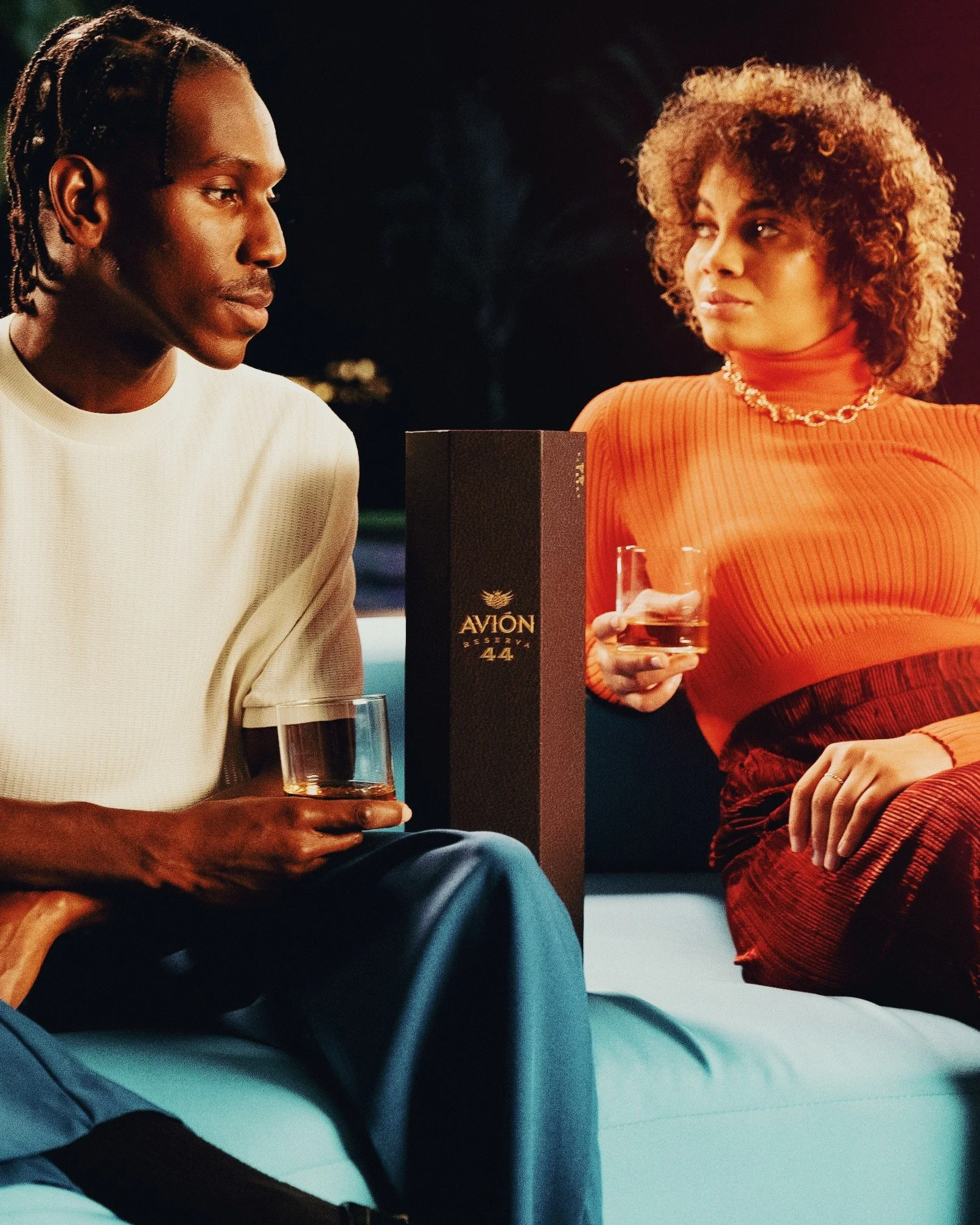 Two women sitting on a couch at night, holding glasses of dark liquor, engaging in conversation. A bottle of Avión tequila is between them.
