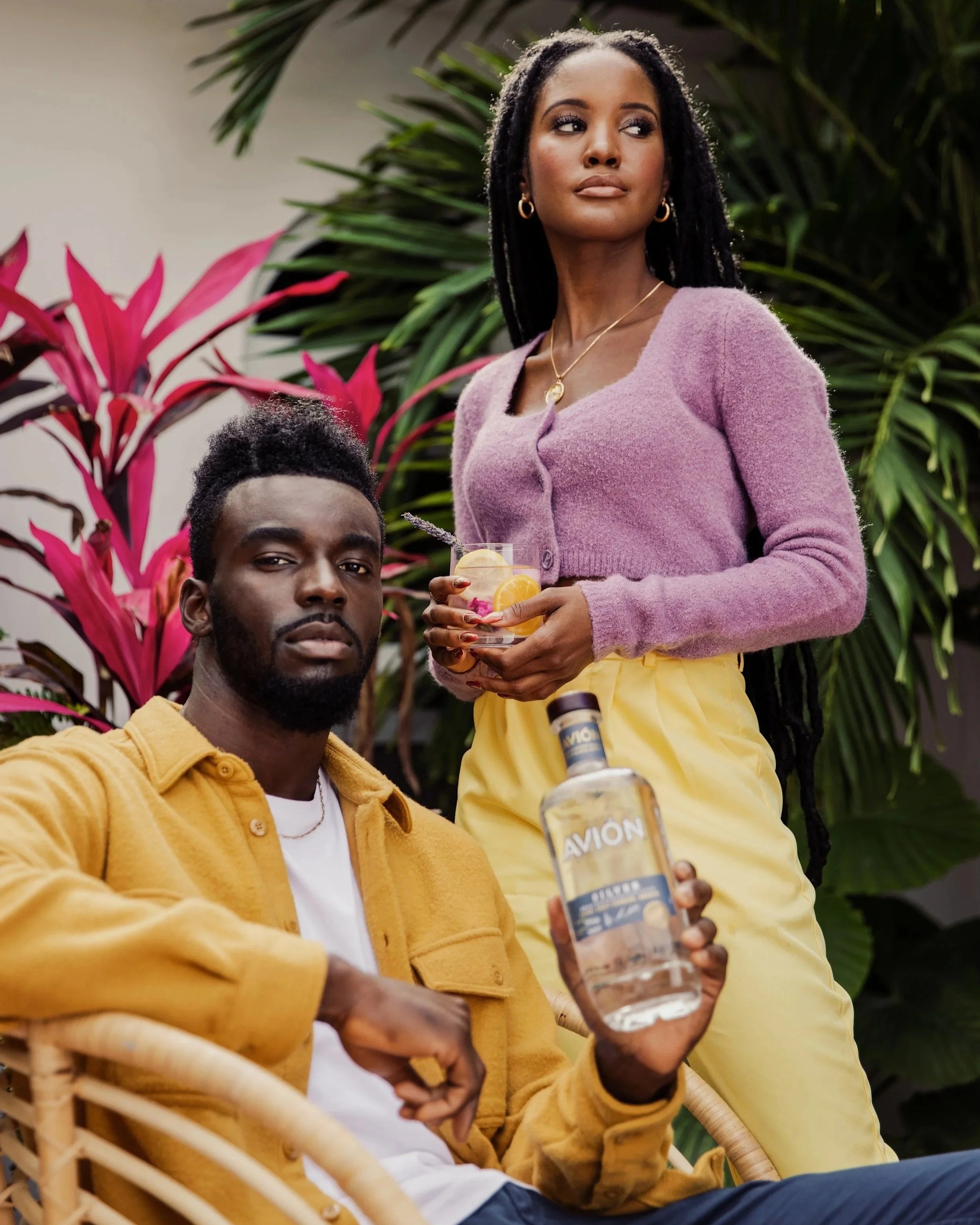 A woman in a pink sweater and yellow pants holding a cocktail, sitting outdoors with a man in a yellow jacket holding an airplane bottle of vodka, surrounded by tropical plants.