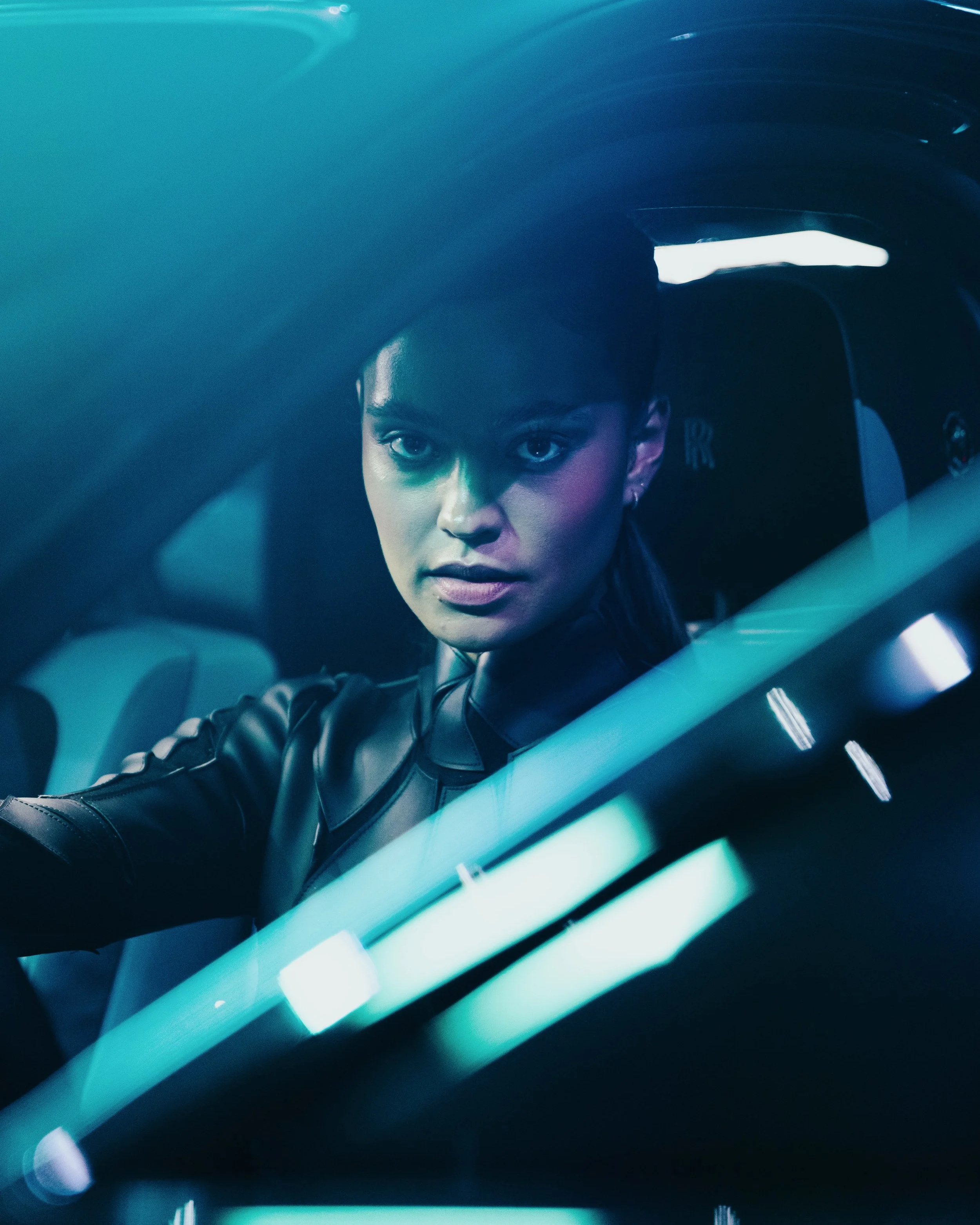 A woman with dark hair and makeup driving a car, with her face partially illuminated by the dashboard lights, seen through the windshield with reflections.