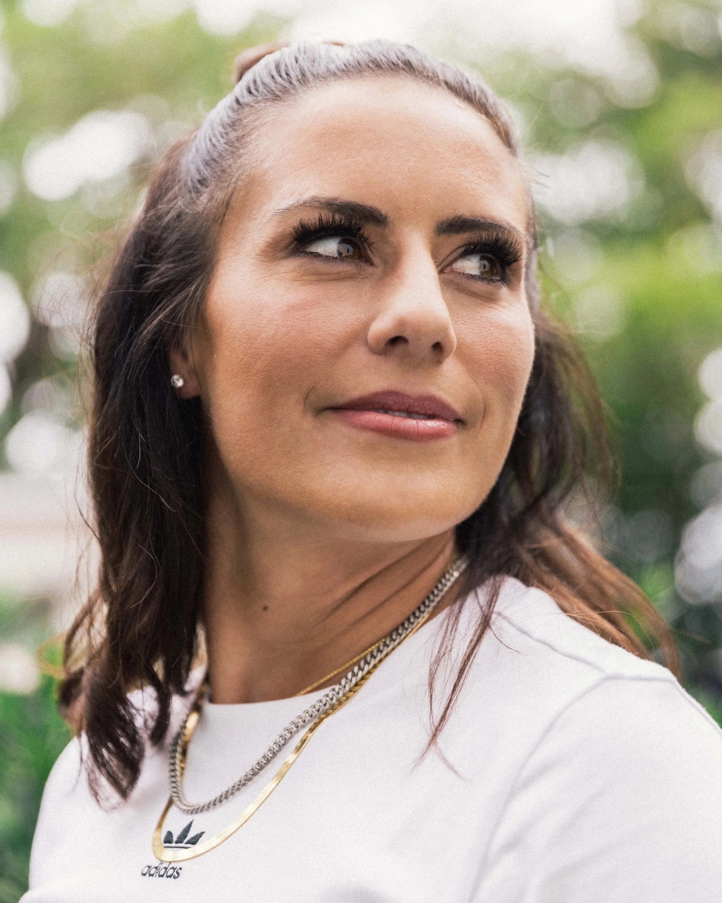 Close-up of a woman with dark hair tied back, wearing jewelry and a white shirt, outdoors with blurred greenery in the background.