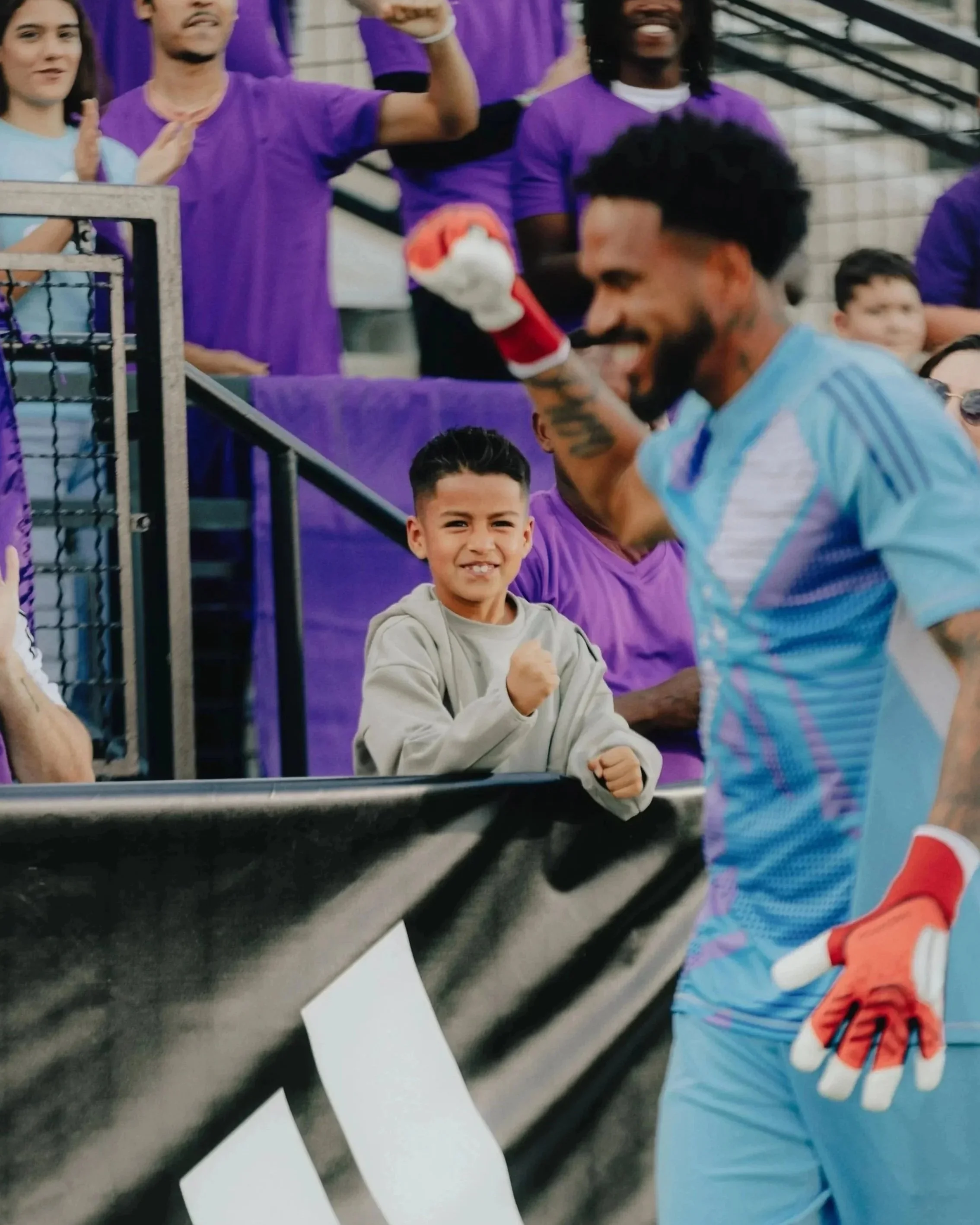 A young boy stands behind a black barrier, smiling and making a fist gesture. A soccer player in a light blue jersey and orange gloves is in the foreground, smiling and gesturing with a fist. Several people wearing purple shirts are in the background
