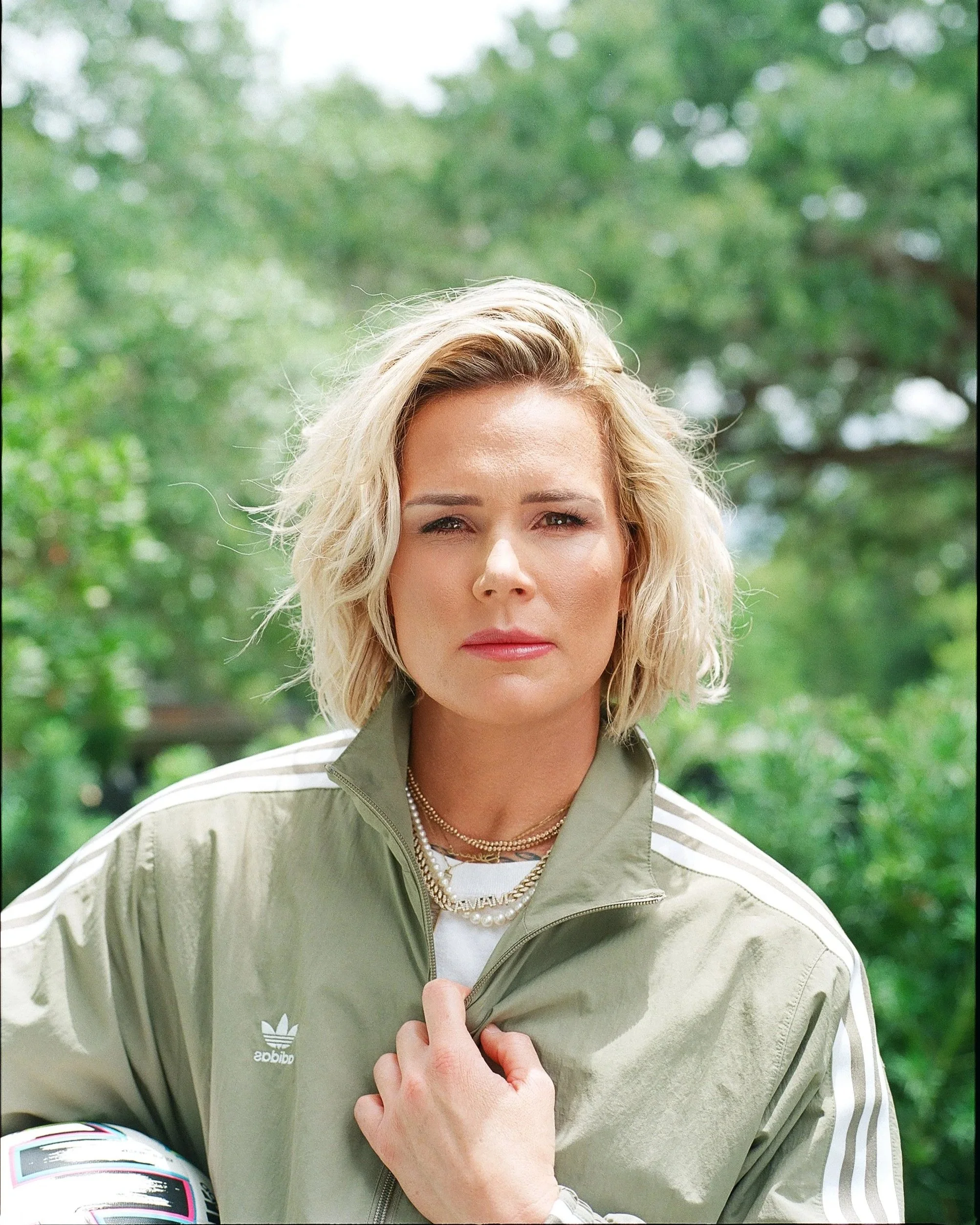 A woman with short blonde hair, wearing a light green Adidas jacket, standing outdoors with green trees in the background.