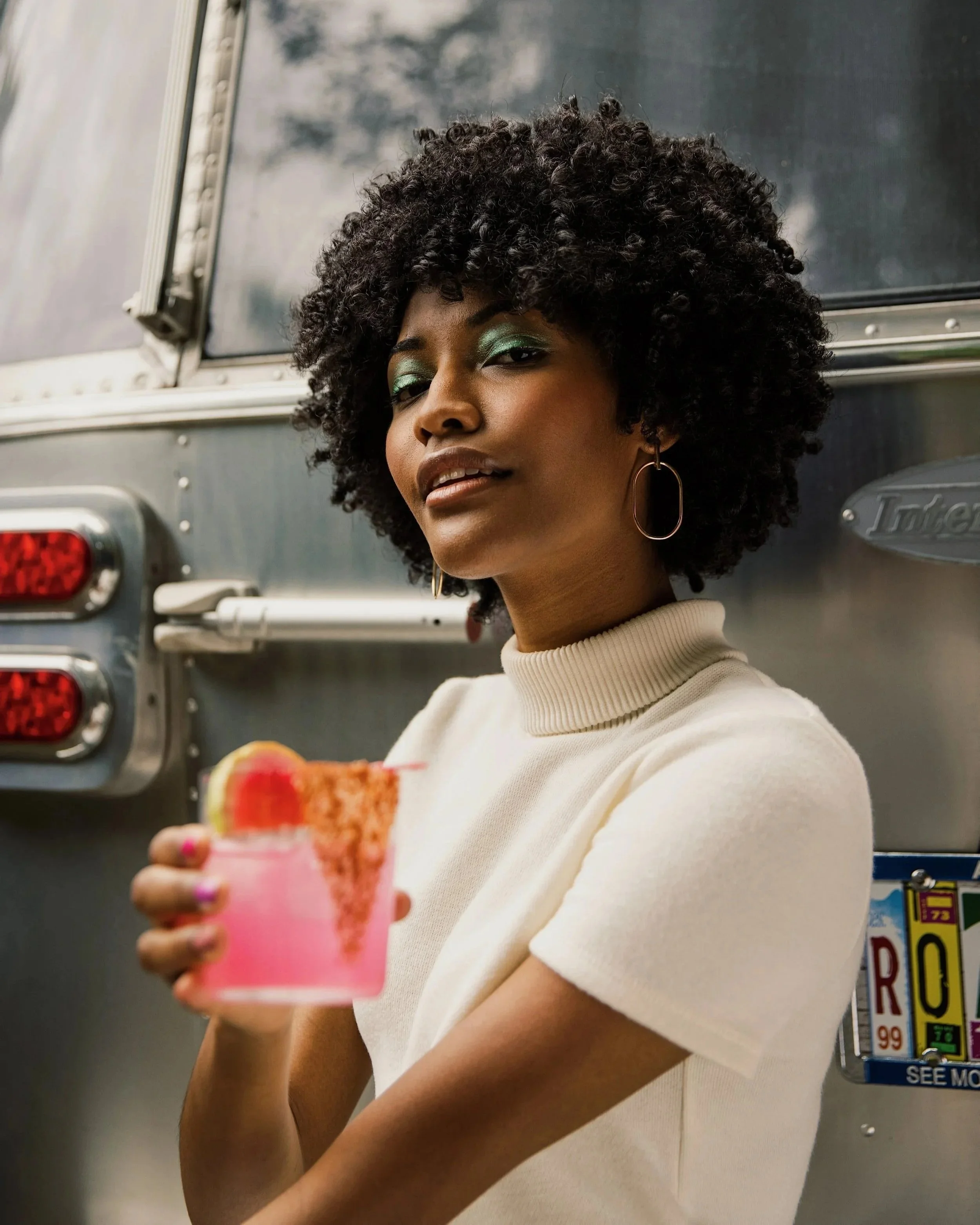 A woman with curly hair and green eye makeup holds a pink cocktail with a slice of lime and a strawberry garnish, standing in front of a metallic food truck or trailer.